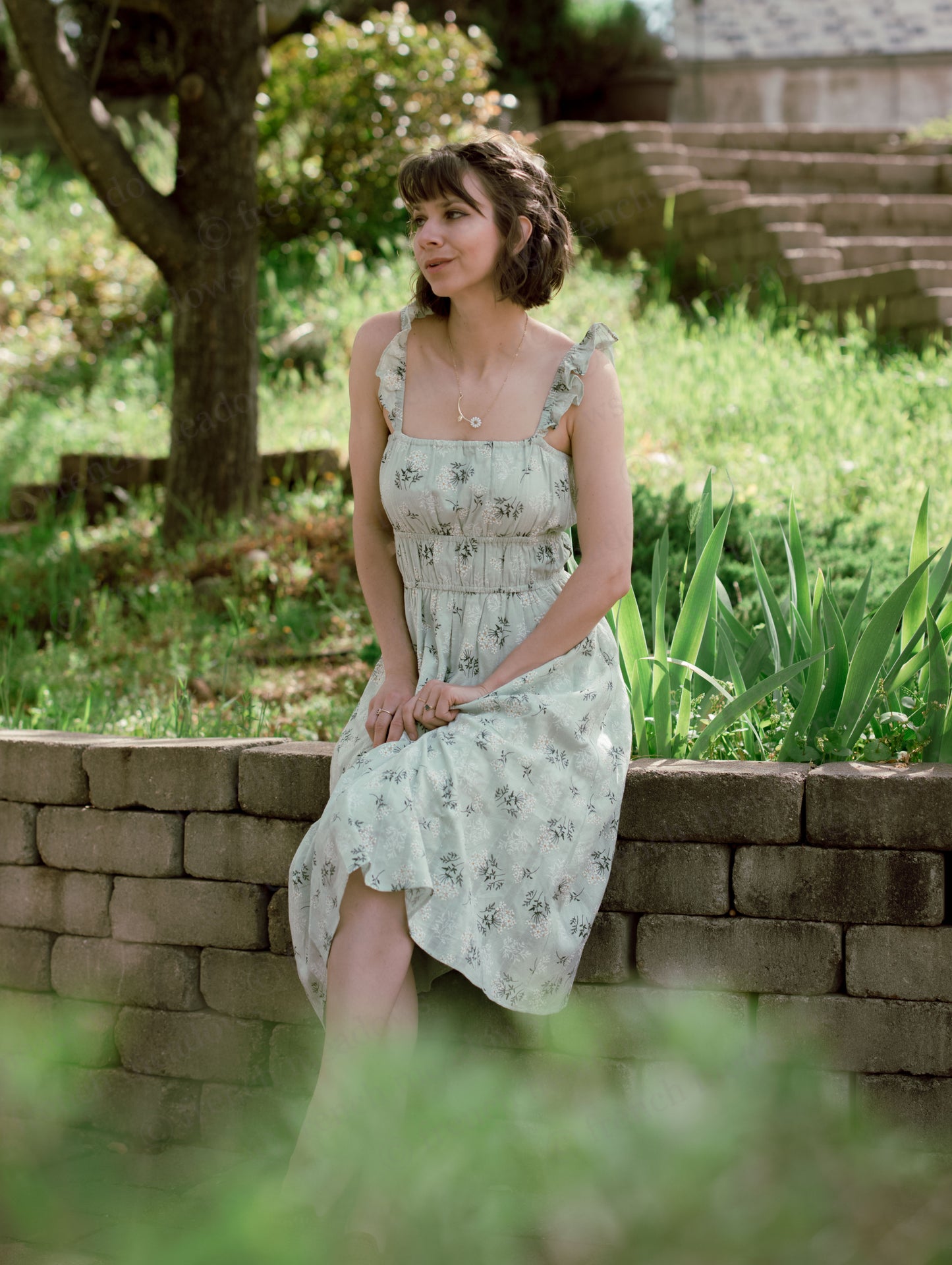 Woman sitting on a stone wall wearing a Floral Milkmaid Dress with ruffled straps and a ruched fitted bodice.