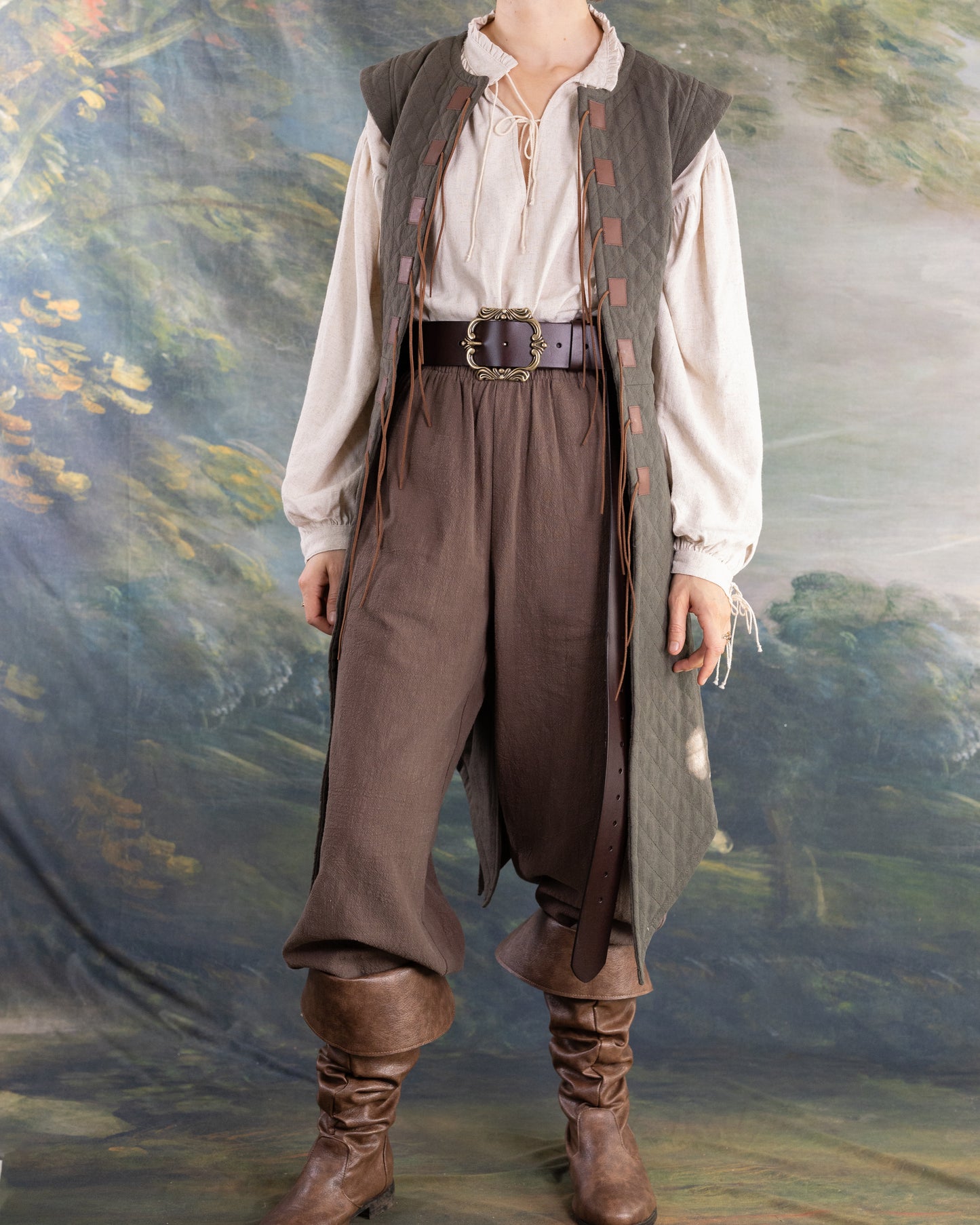 Woman wearing a long brown Quilted Gambeson over cream blouse and pants with tall leather boots against painted backdrop