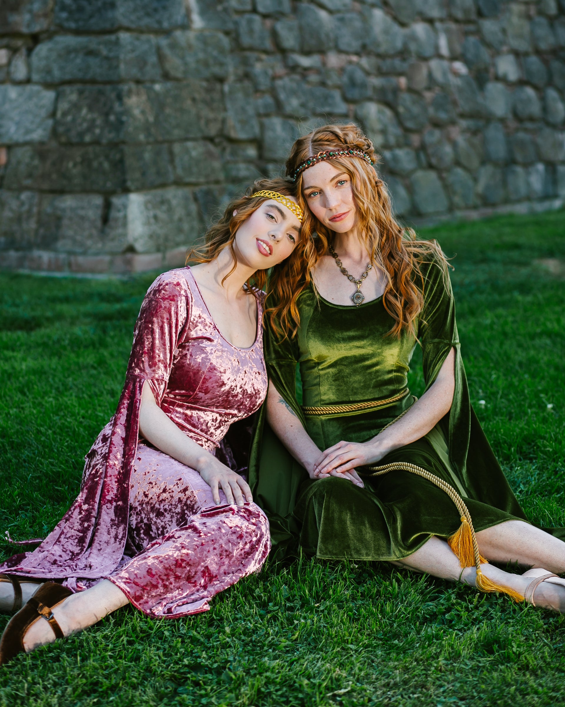 Two women sitting on grass wearing velvet medieval-style dresses with long bell sleeves, one in chartreuse green and one in pink