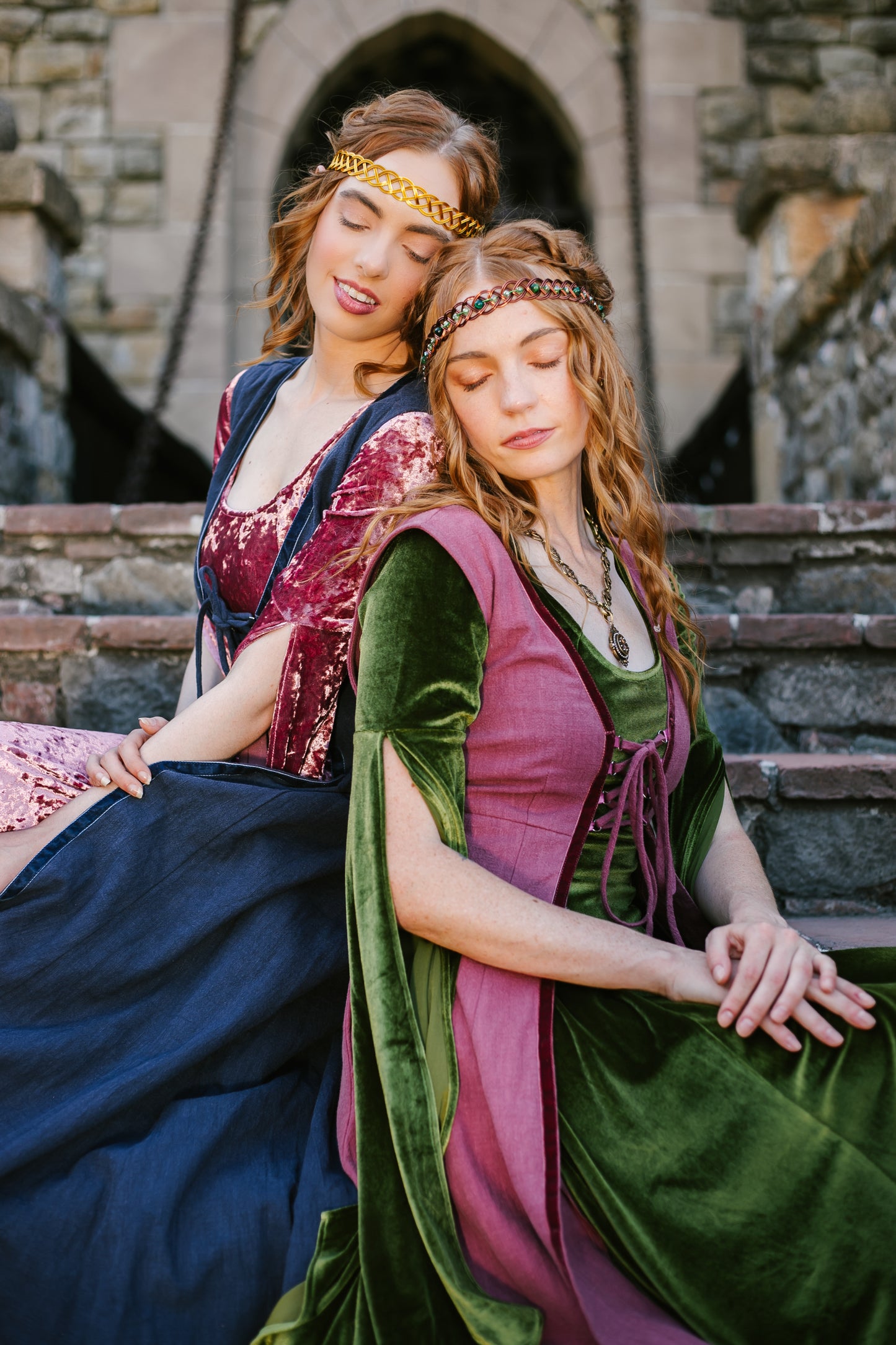 Two women wearing medieval style velvet dresses with bell sleeves sitting on stone steps by an arched doorway