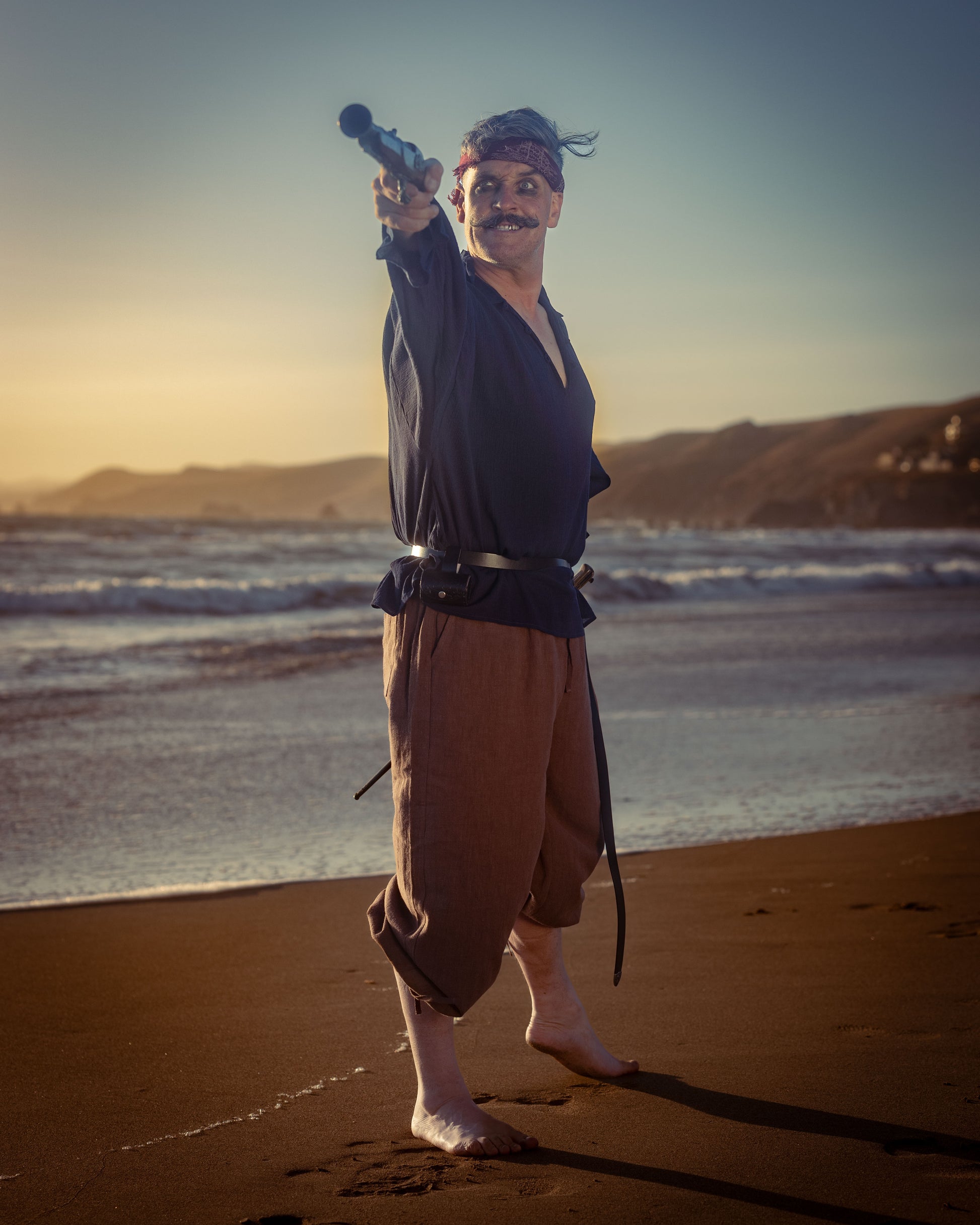 Man wearing rustic pirate pants and shirt pointing a telescope at the beach during sunset