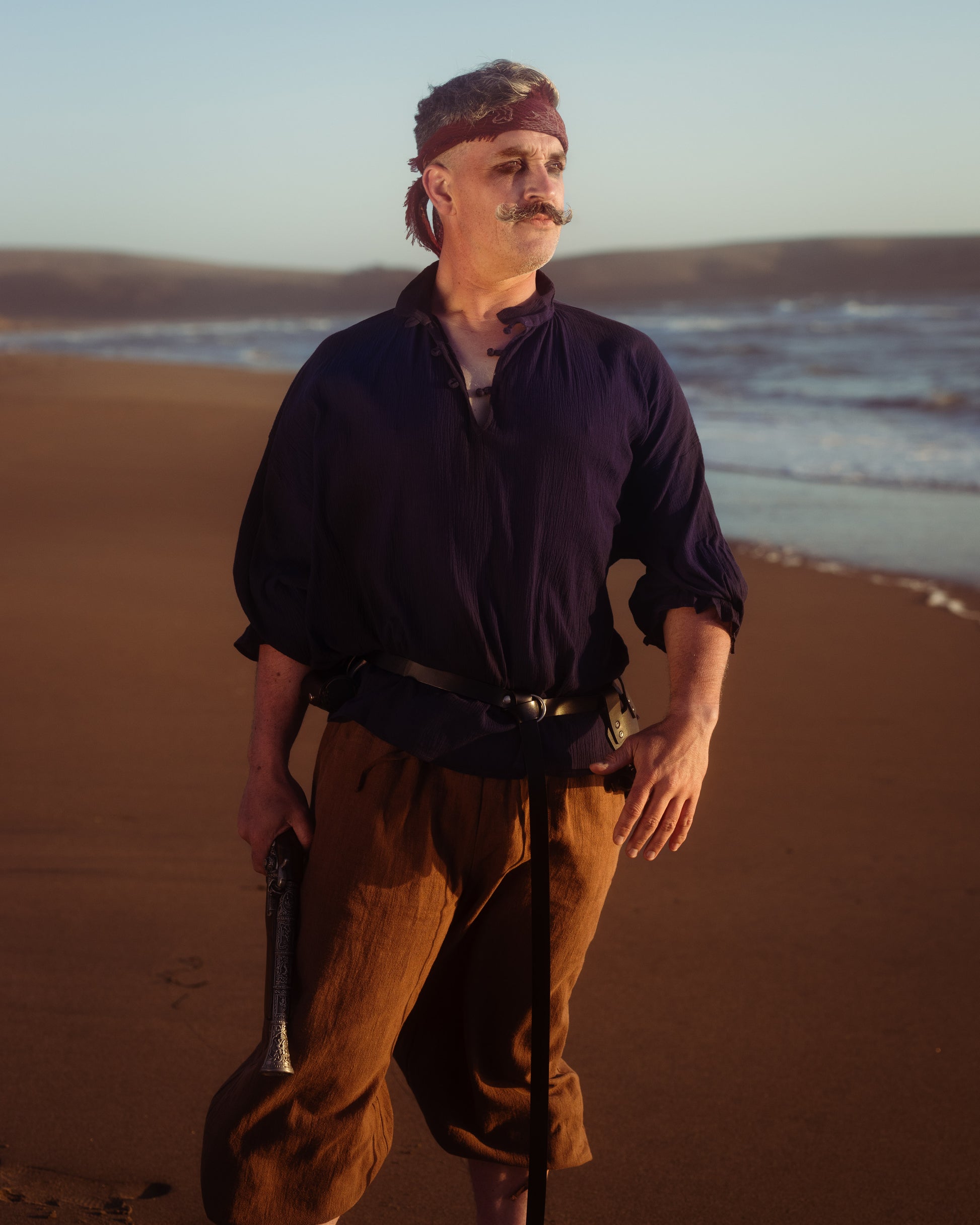 Man wearing a dark billowing poet shirt, brown pants, and a headband standing on a beach at sunset.