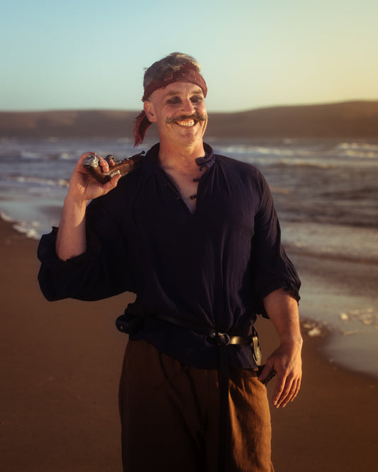 Man wearing a dark billowing poet shirt with knotted buttons, holding a pistol on a beach at sunset.