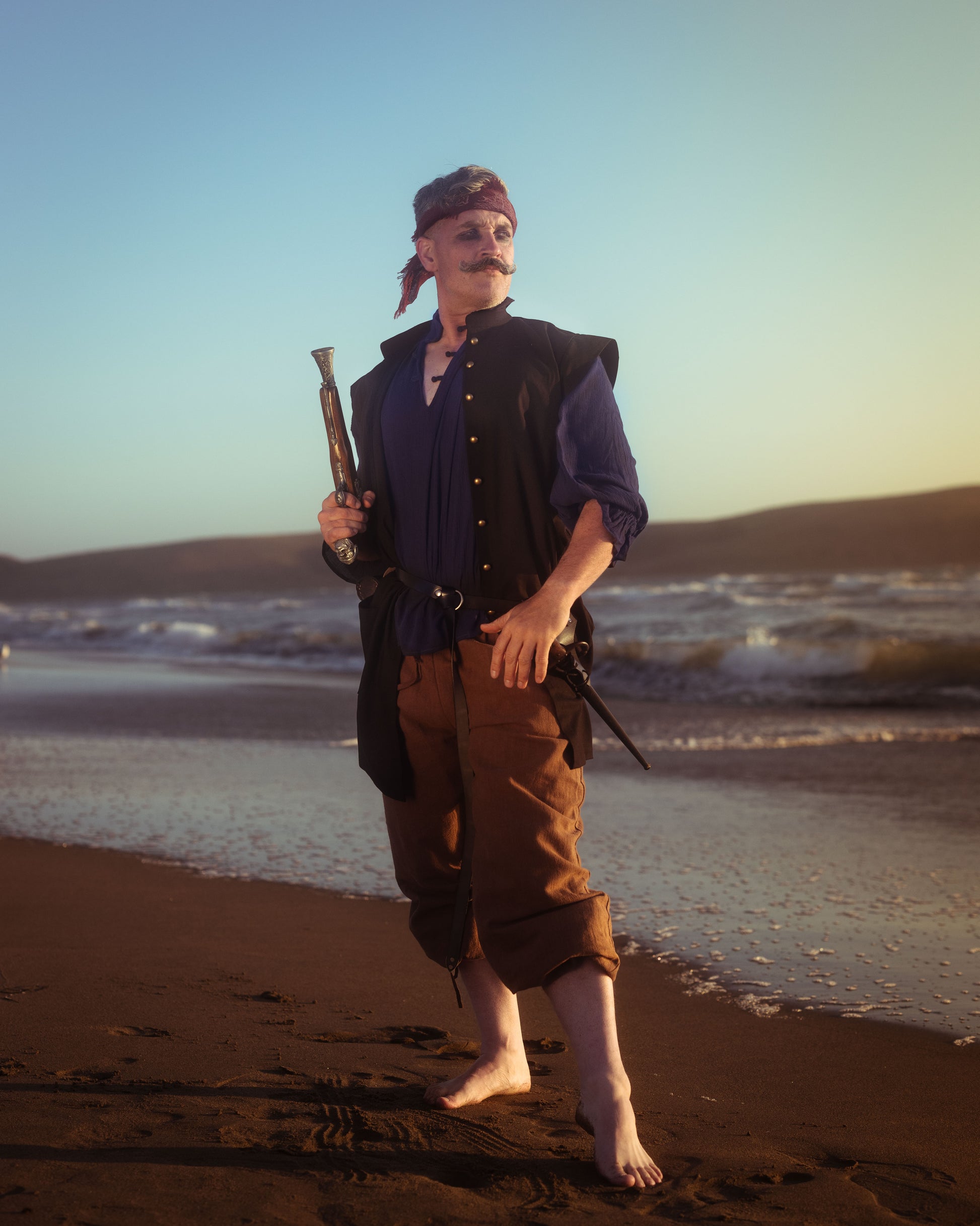 Man wearing a black pirate vest over a blue shirt with brown pants holding a spyglass on a beach at sunset