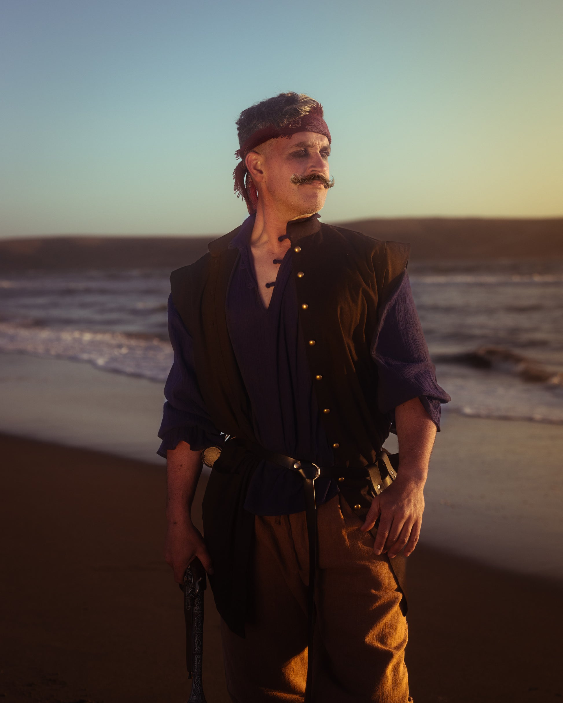 Man wearing black pirate vest over blue shirt, standing on beach at sunset, styled as historical pirate costume