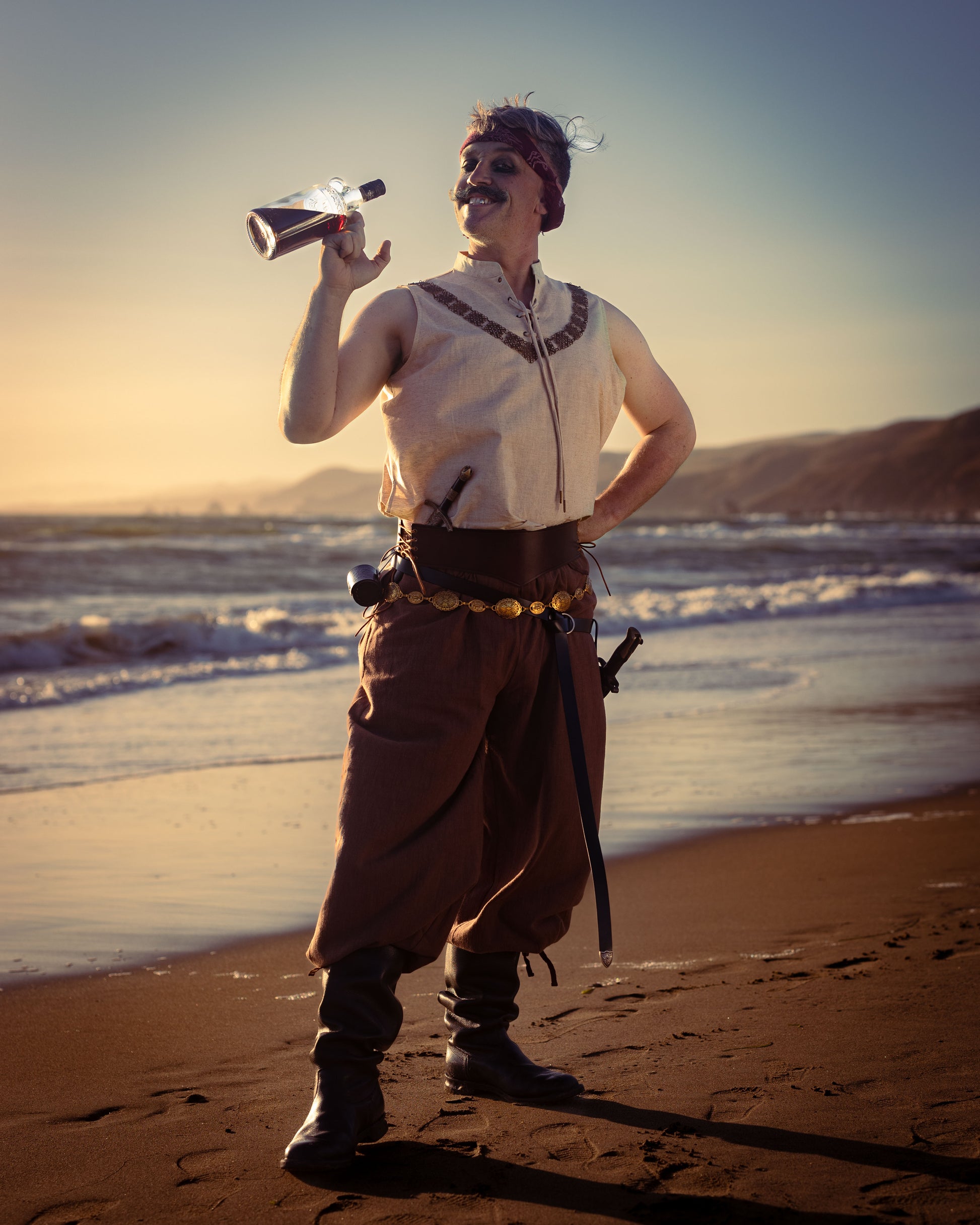 Man wearing rustic wide-leg brown pirate pants with drawstring waist standing on beach at sunset