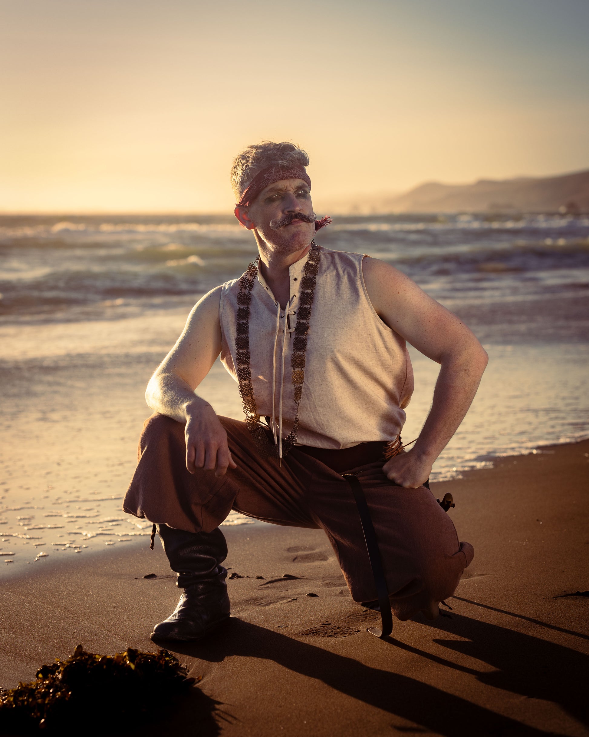 Man wearing rustic wide leg pirate pants with boots and sleeveless shirt posing on beach at sunset
