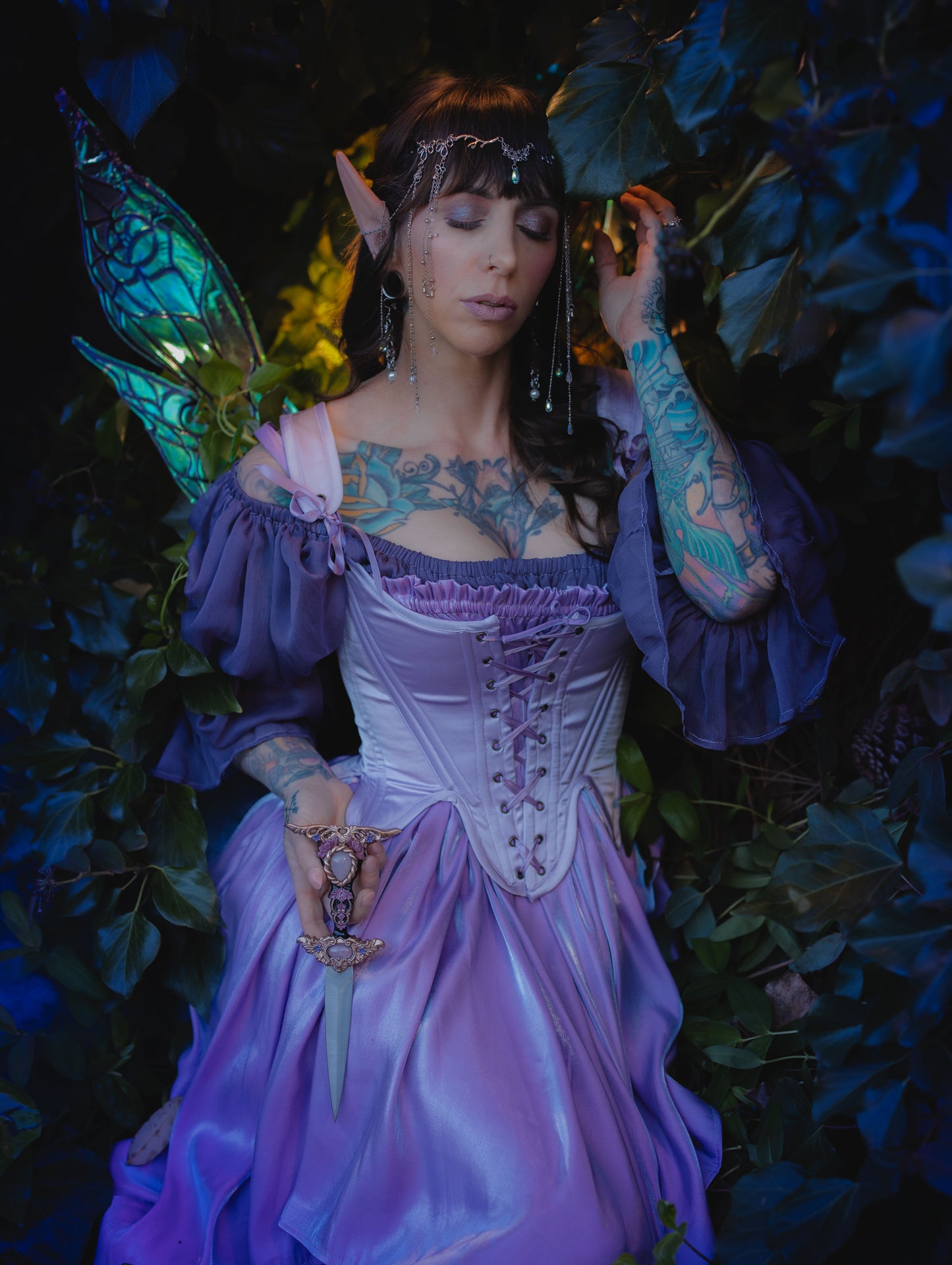 Woman in Lilac Satin 18th Century Stays with elf ears and fairy wings in a dark botanical setting.