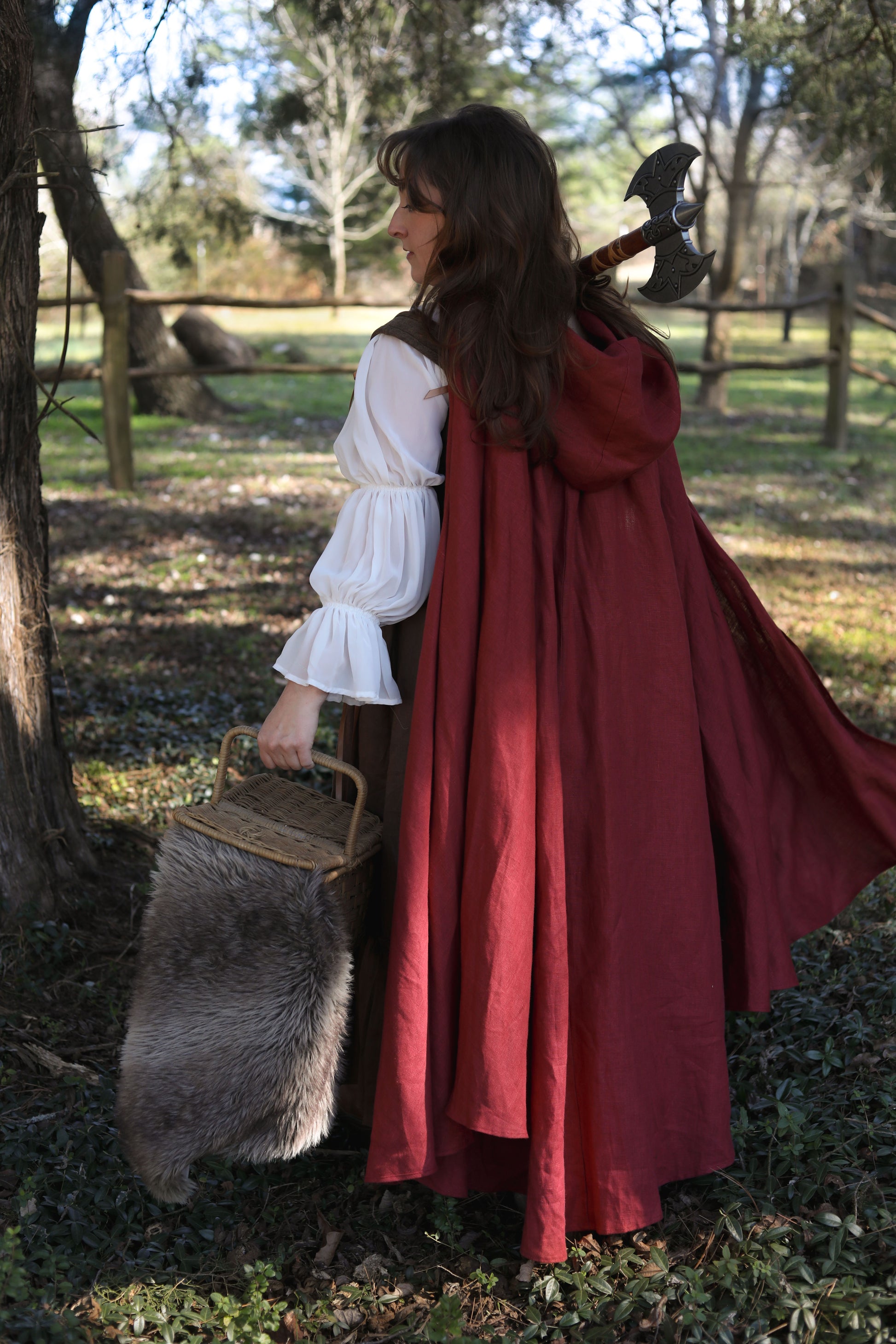 Woman wearing a deep red linen renaissance cloak with hood, carrying a basket and standing in a forest setting by a wooden fence.