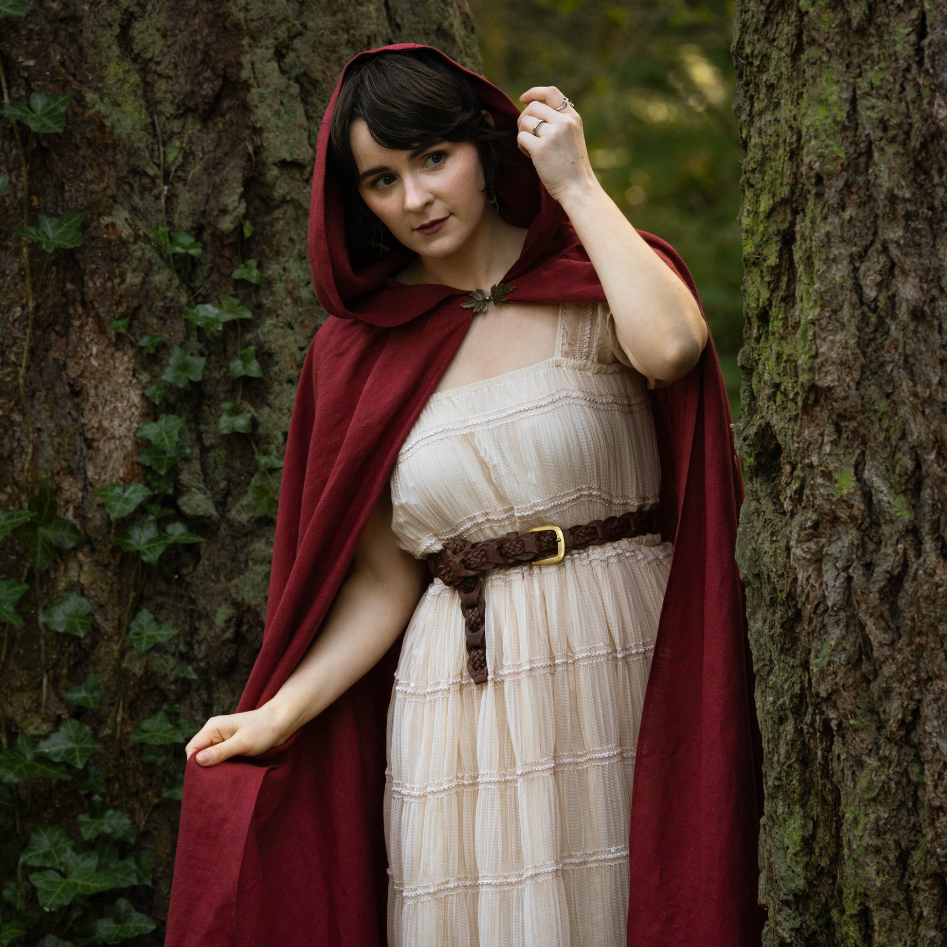 Woman wearing a deep red Linen Renaissance Cloak over a cream dress standing between two large trees in a forest setting