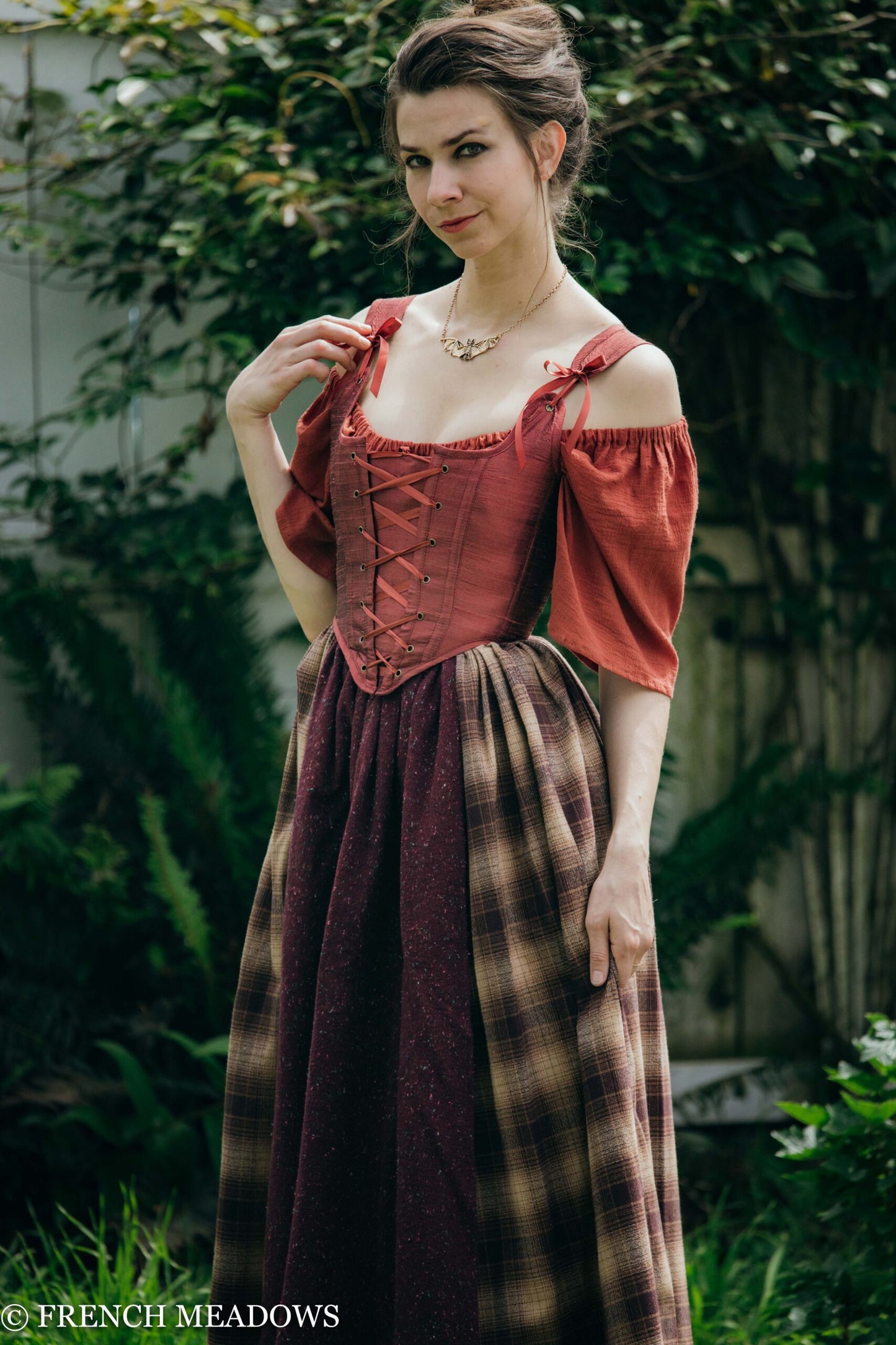 Mary Sanderson Skirt in Brown Plaid shown with red corset and off-shoulder sleeves in garden setting against greenery