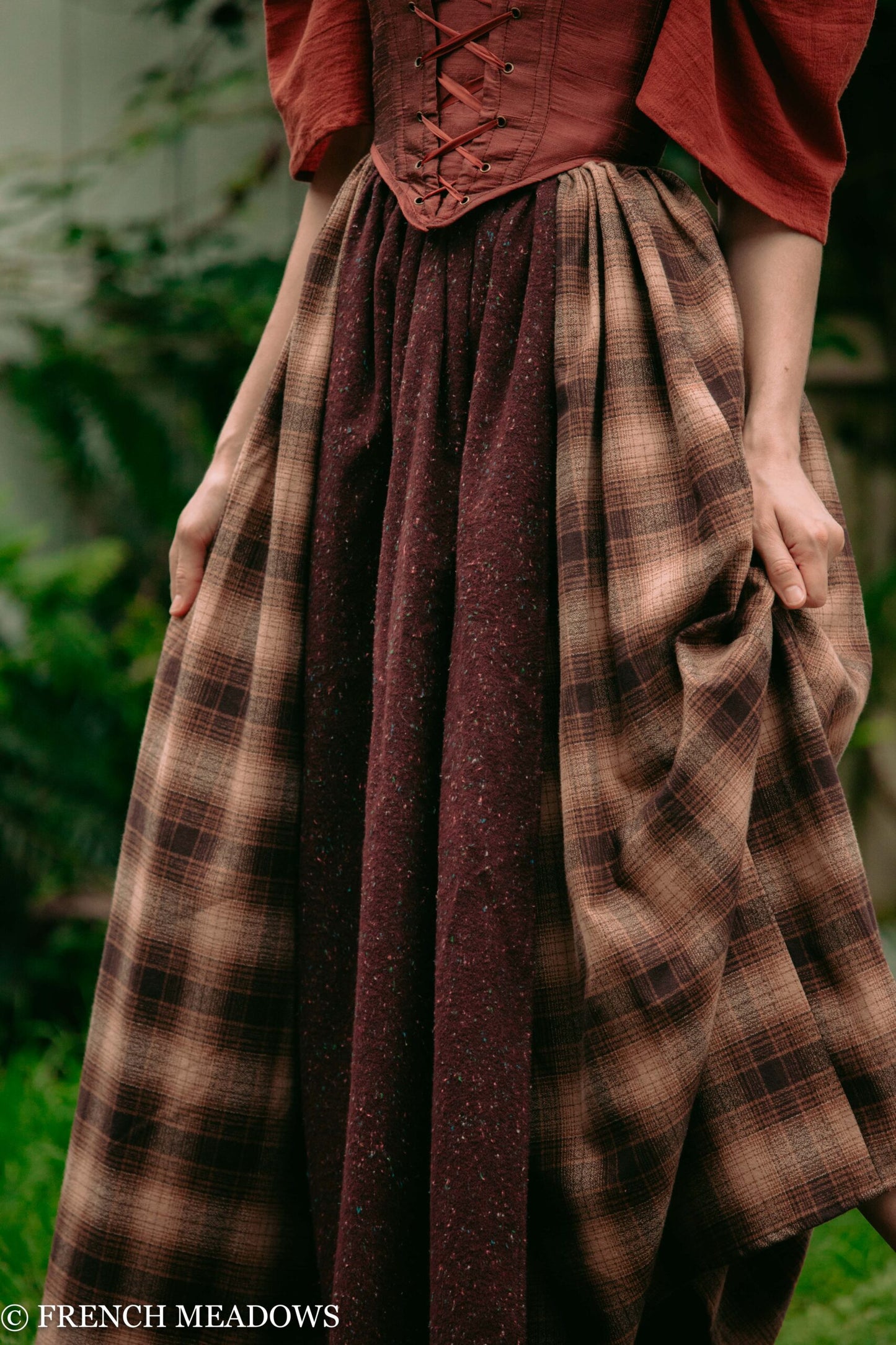Mary Sanderson Skirt in brown plaid cotton with purple apron overlay, shown with red corset against natural green background