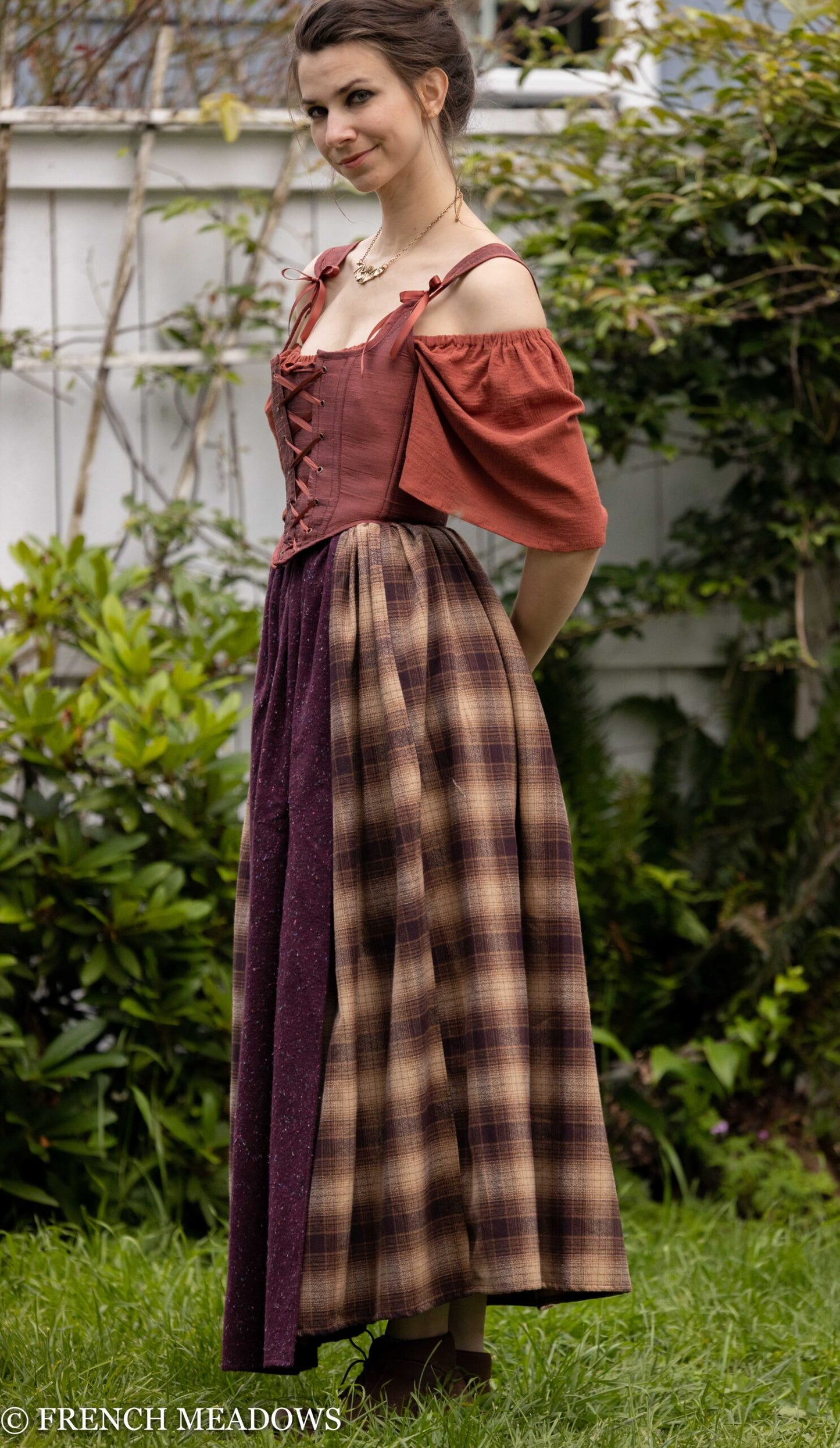 Mary Sanderson Skirt in Brown Plaid features plaid cotton skirt with purple apron overlay, shown outdoors against garden backdrop