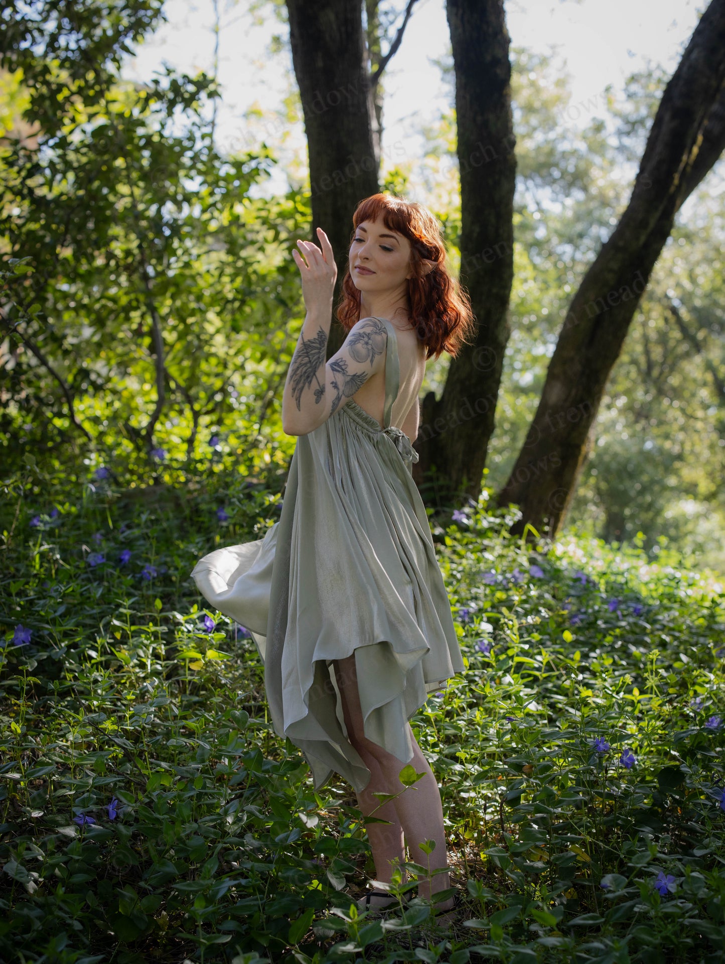 Woman with red hair wearing a flowing Flutter Fairy Dress with handkerchief hem in a sunlit forest garden.