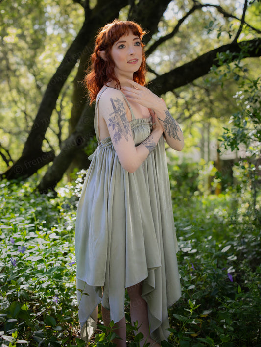 Woman wearing a flowing Flutter Fairy Dress with handkerchief hem standing in a sunlit forest clearing.