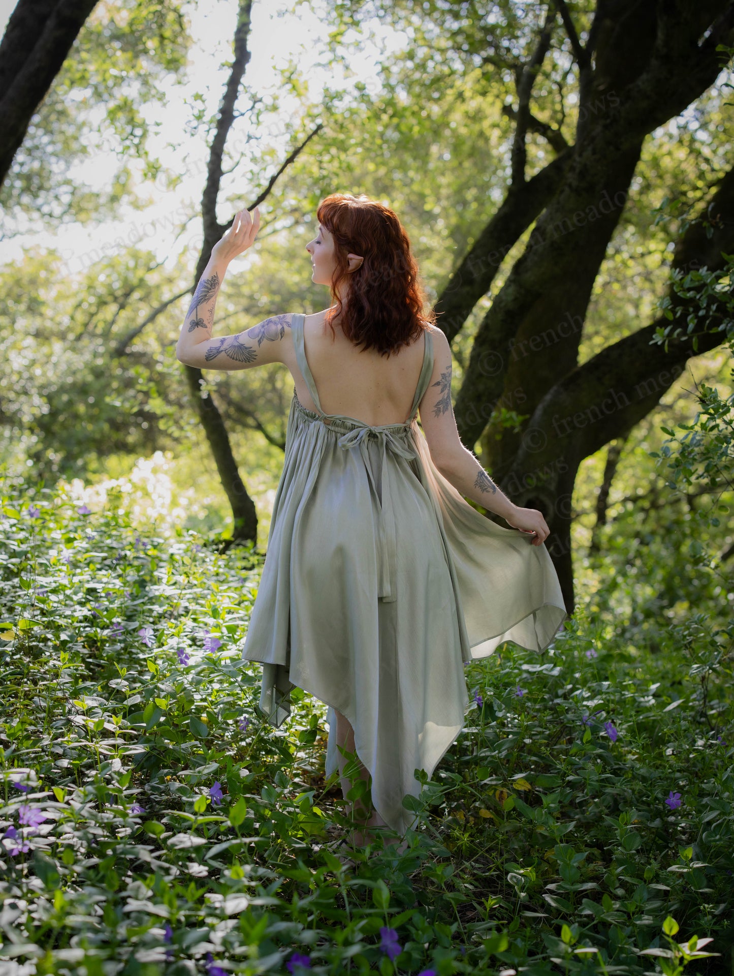 Woman in a flowy pale green Flutter Fairy Dress with low back and handkerchief hem in a sunlit garden setting
