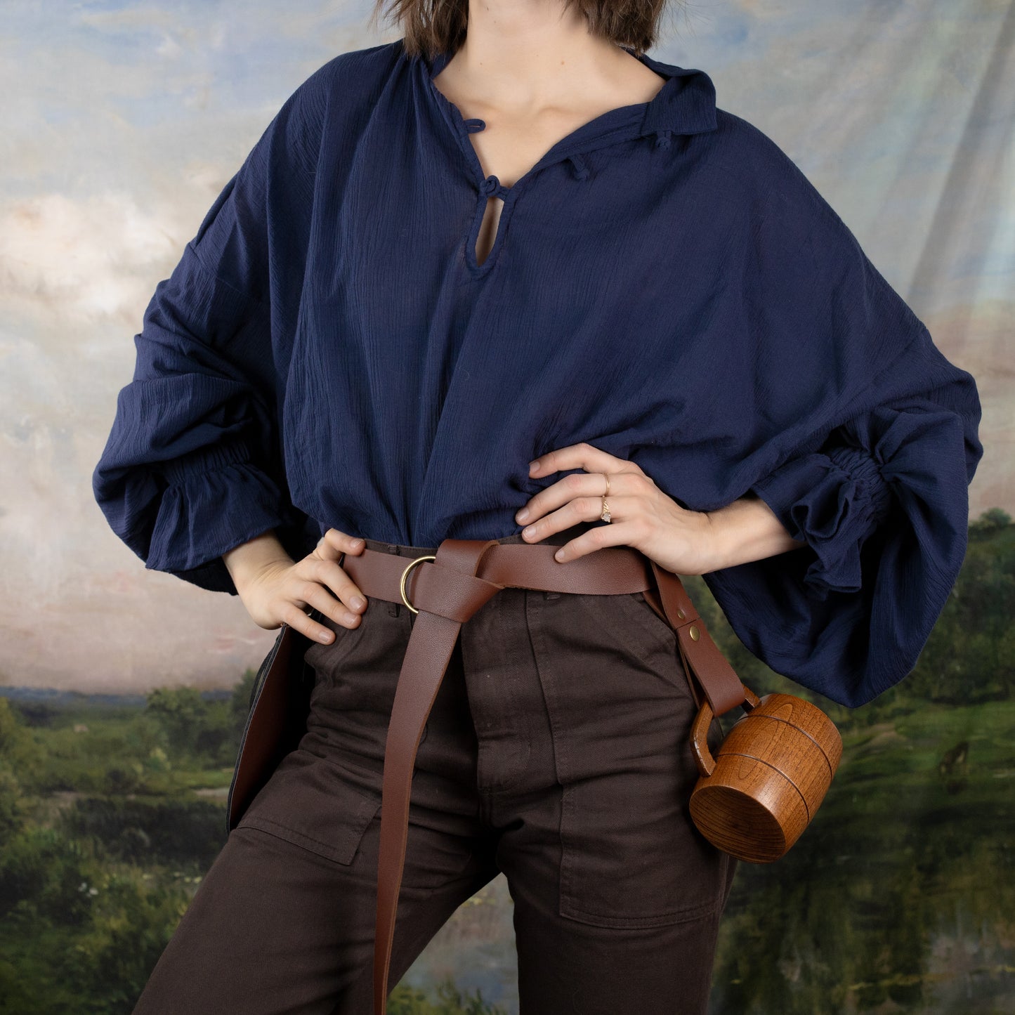 Darcy Shirt in navy blue cotton with billowing sleeves, brown leather belt, and rustic tankard against pastoral backdrop