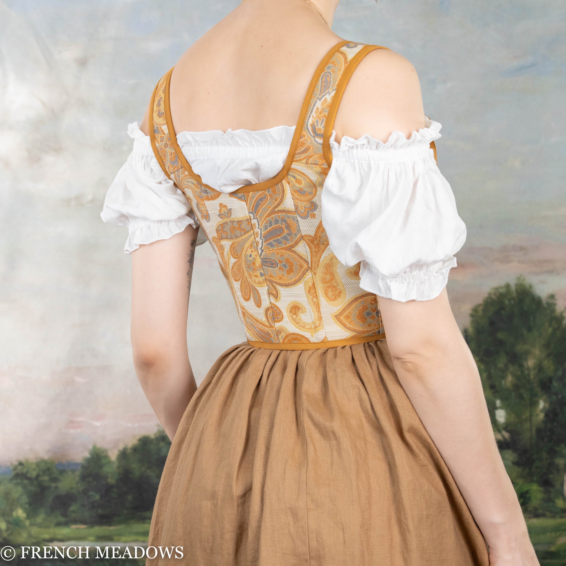 Orange and yellow paisley Renaissance bodice with gold trim and white puff sleeves shown from back against pastoral backdrop
