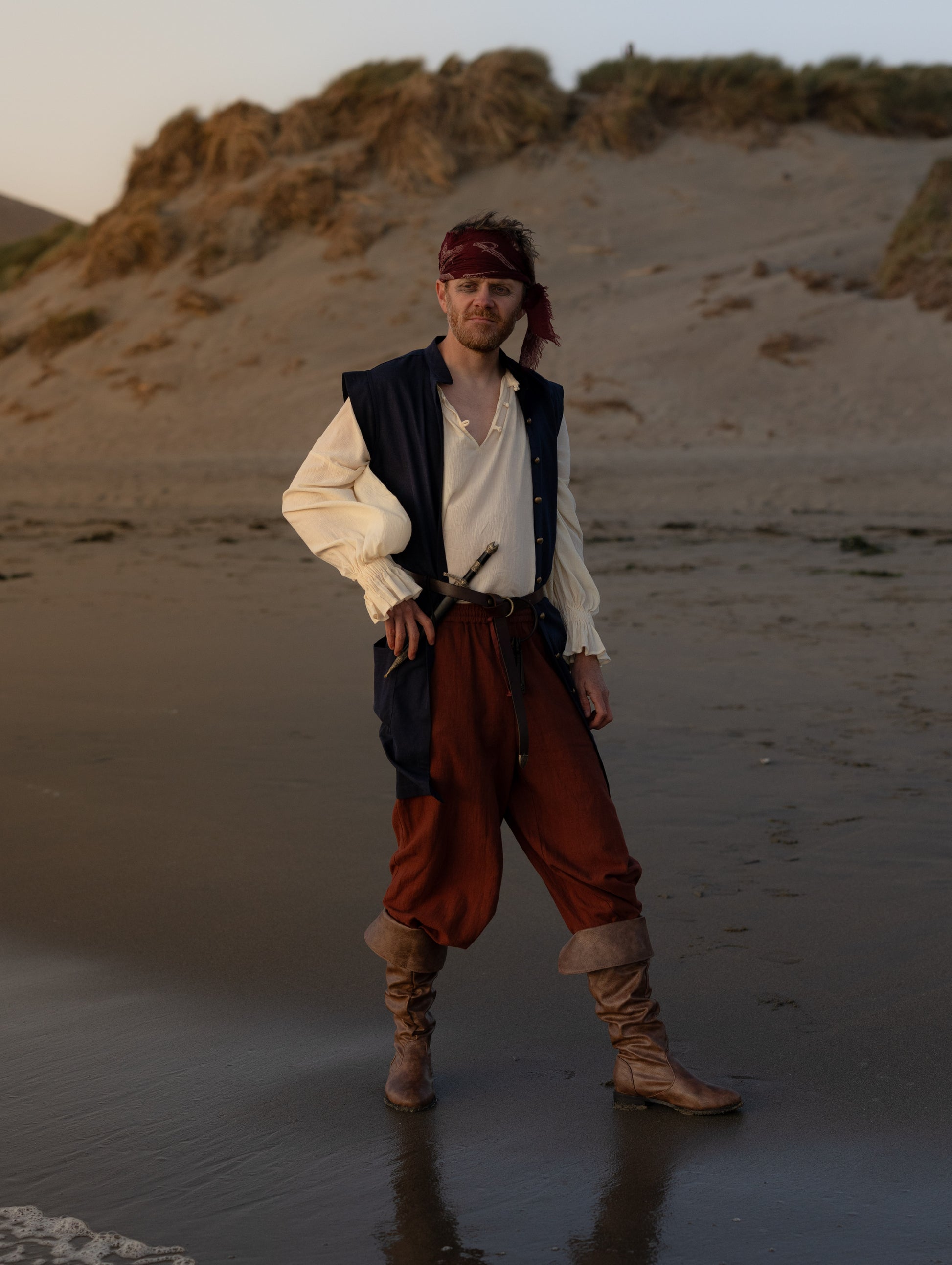 Man wearing rustic wide-leg pirate pants with boots and a vest standing on a beach at sunset