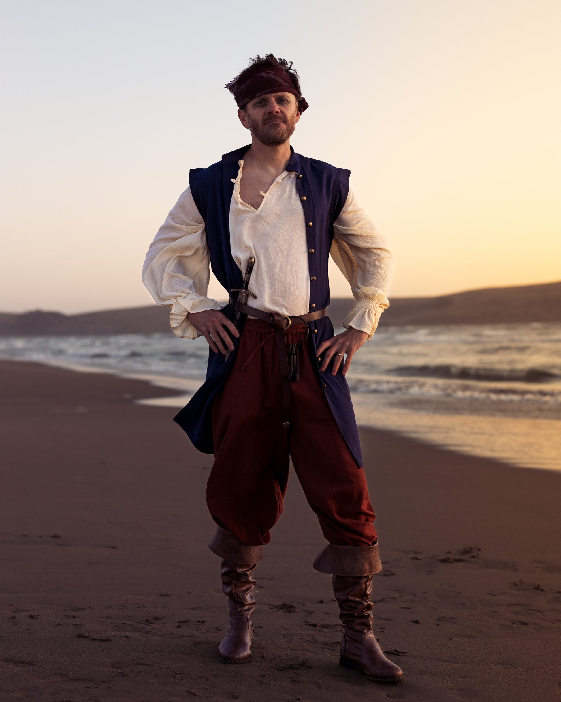 Man wearing rustic pirate pants with wide legs and drawstring waist standing on beach at sunset