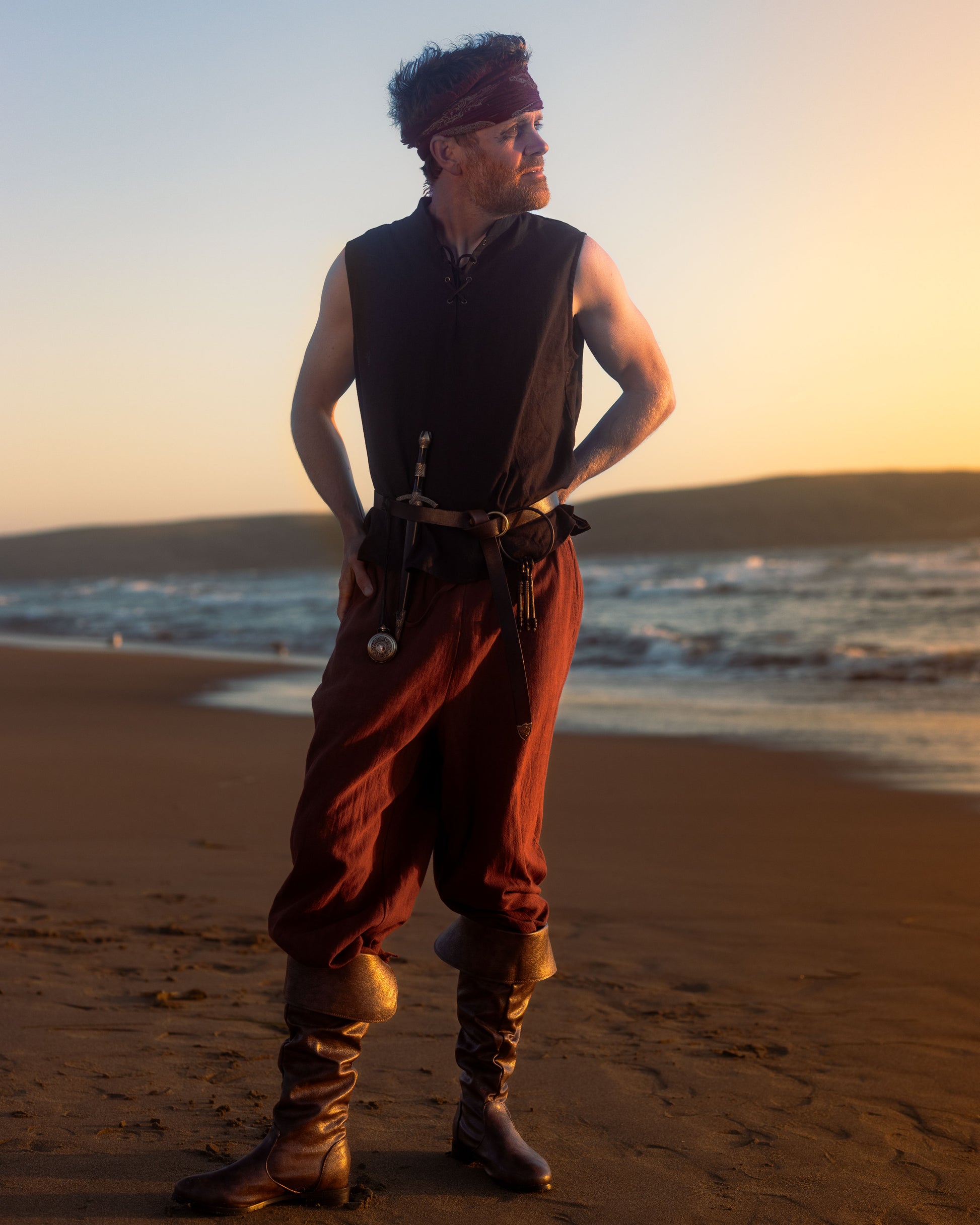 Man wearing rustic wide-leg pirate pants with boots and sleeveless vest standing on a beach at sunset