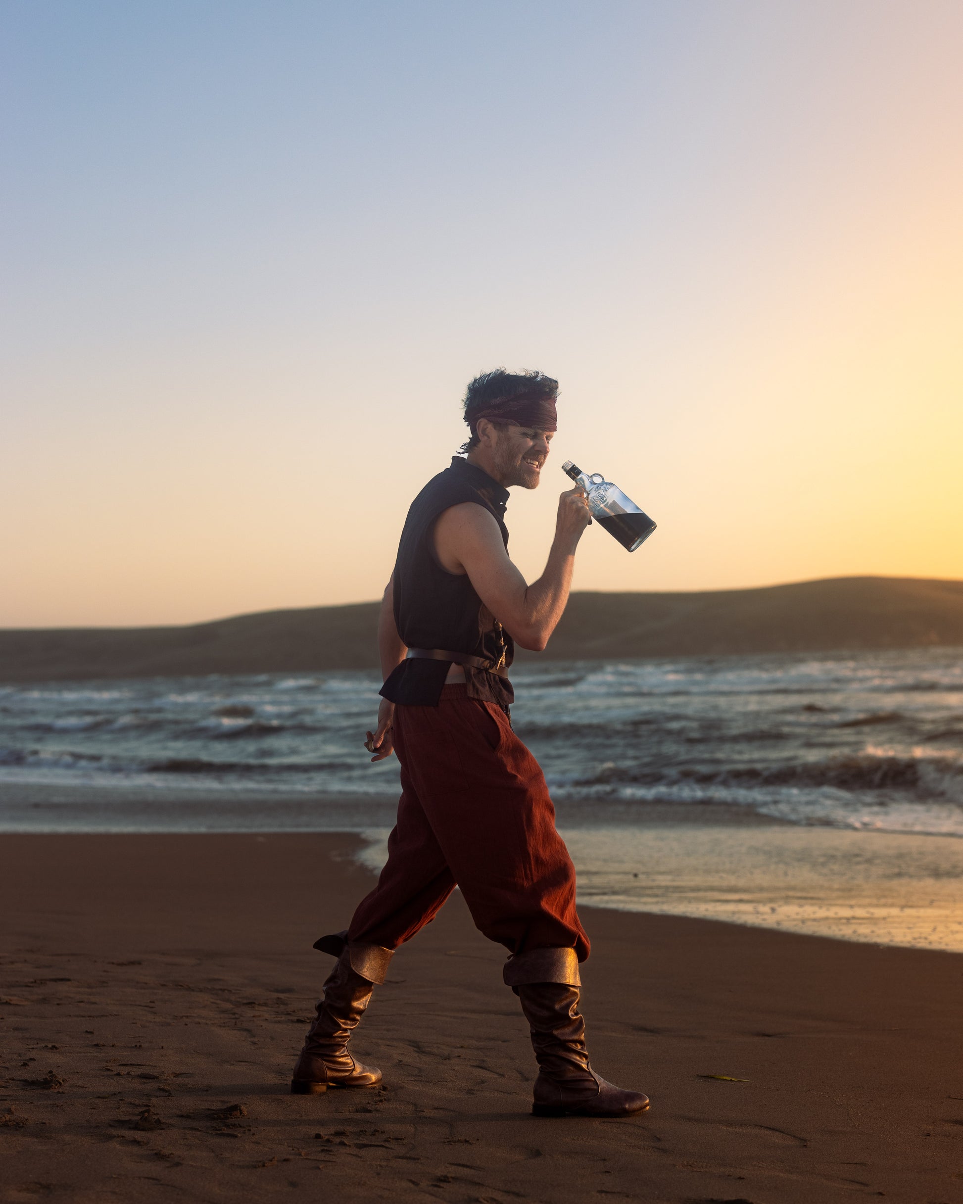 Man wearing rustic wide-leg pirate pants walking on beach at sunset holding a bottle and dressed in boots and sleeveless shirt