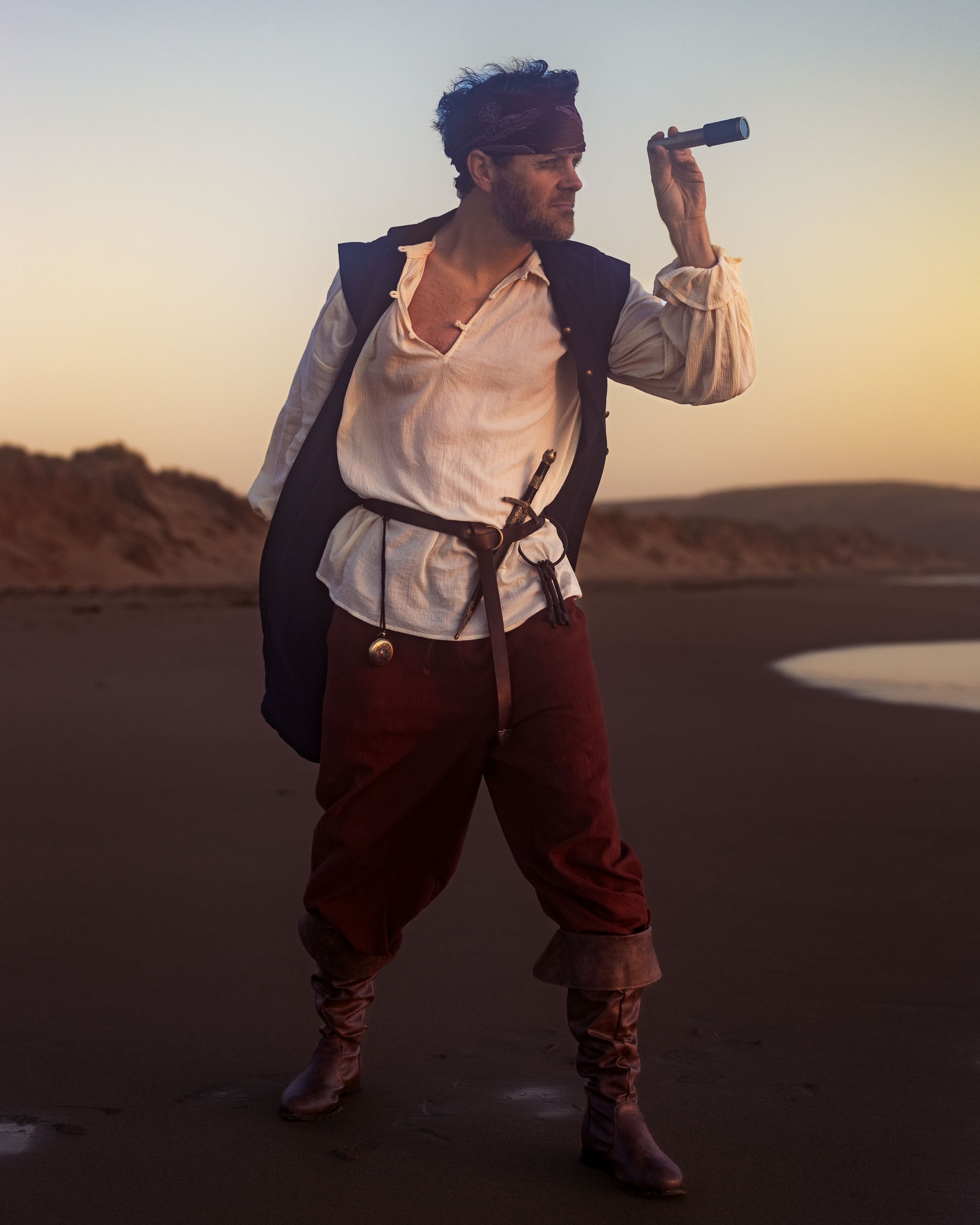 Man wearing antique ivory Darcy Shirt with billowing sleeves and knotted buttons, looking through a spyglass at sunset.