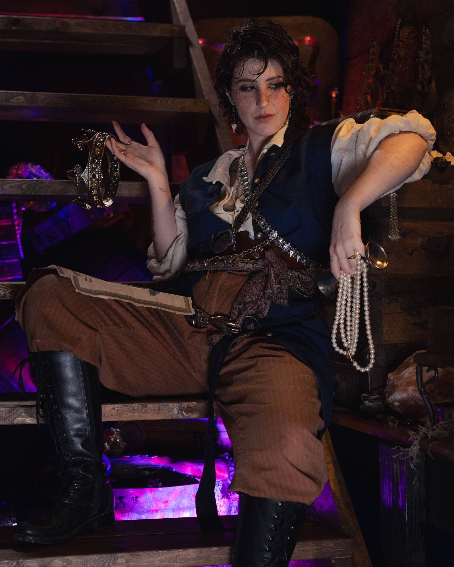 Navy Pirate Vest shown with pearl necklace and tambourine in atmospheric purple-lit setting with wooden beams and brown pants