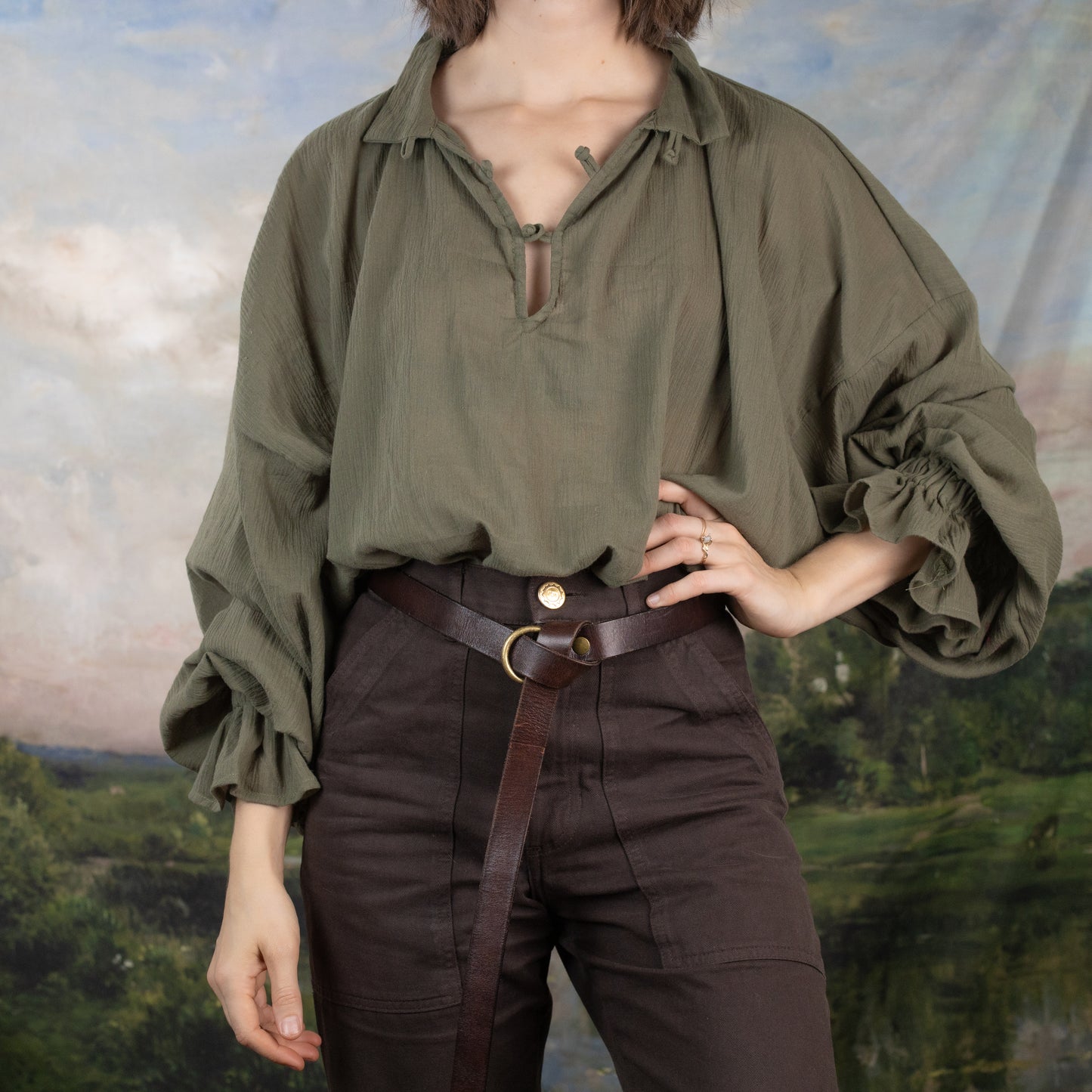 Darcy Shirt in olive green cotton with billowing sleeves and knotted neckline, styled with brown belted pants against pastoral backdrop