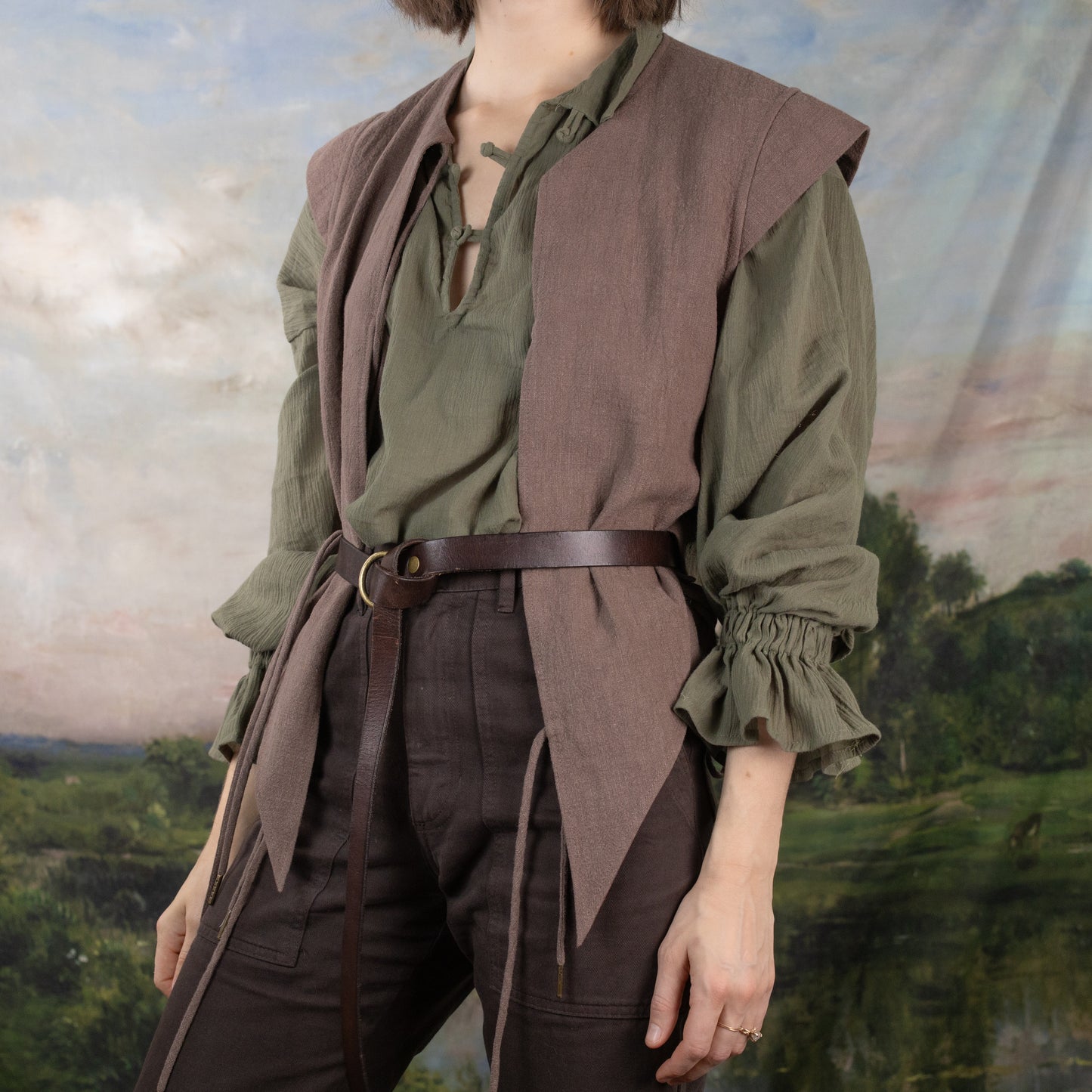 Darcy Shirt styled in sage green with billowing sleeves, paired with brown vest and leather belt against pastoral backdrop