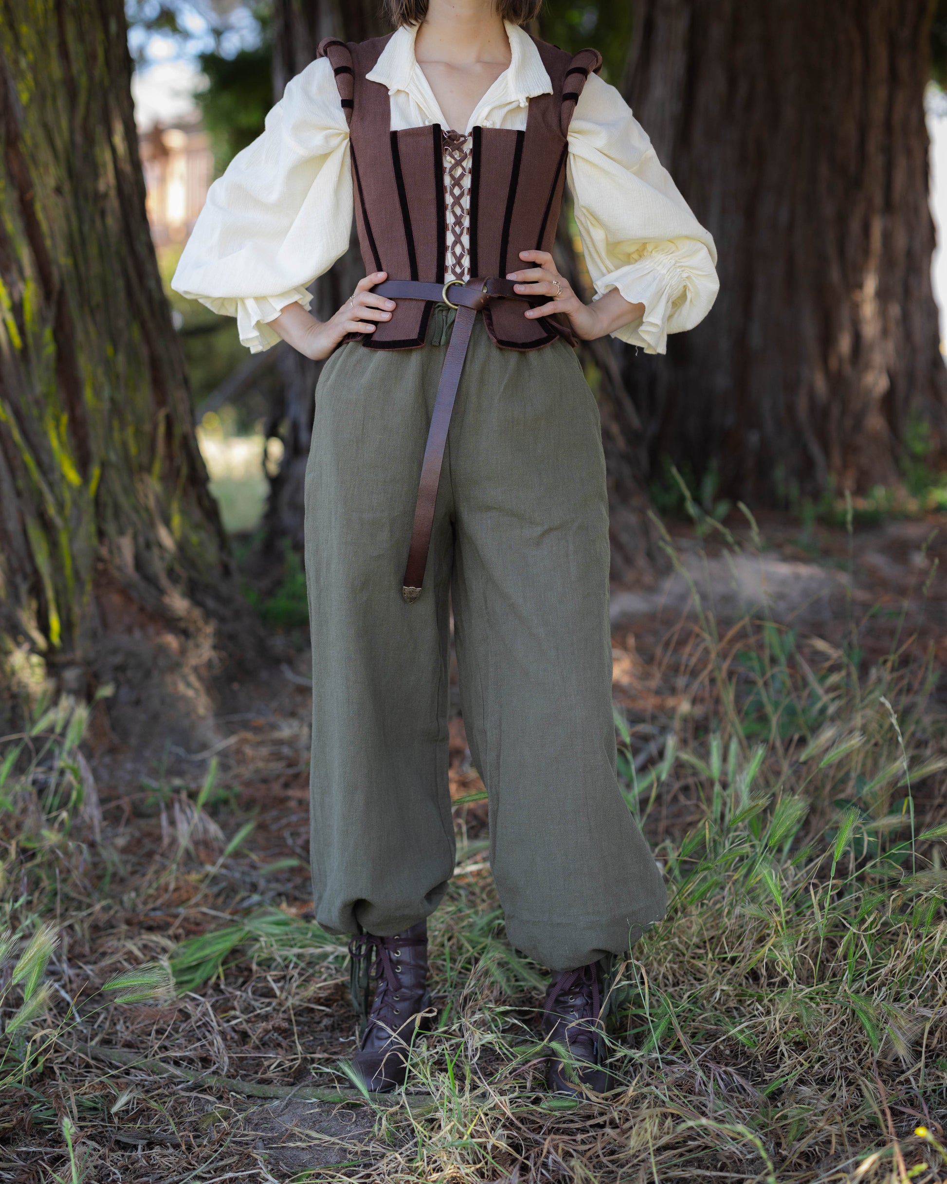 Person wearing a brown Tavern Bodice with lace-up front over a cream blouse and loose green pants in a forest setting.