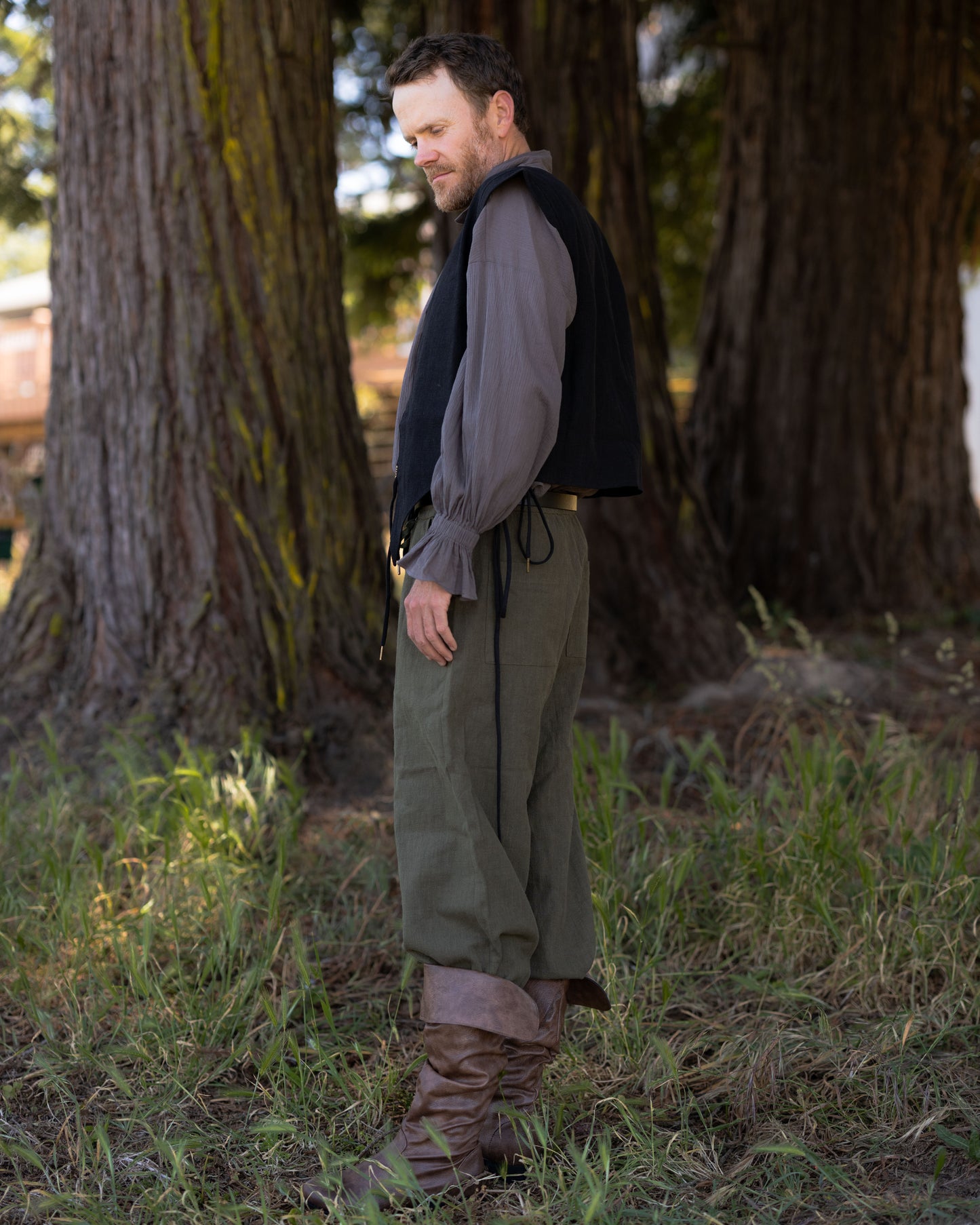 Man wearing Black Bard Vest with gray shirt and green pants standing outdoors by large trees