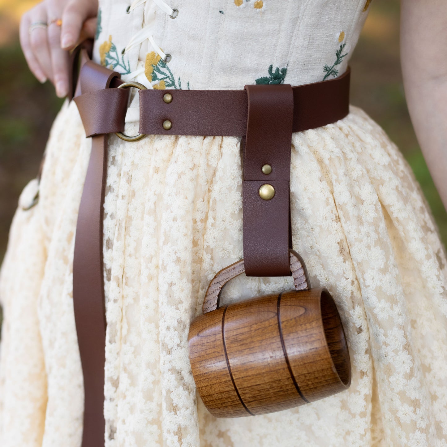Vegan leather tankard strap in brown with metal snap attached to belt, holding wooden mug against floral dress