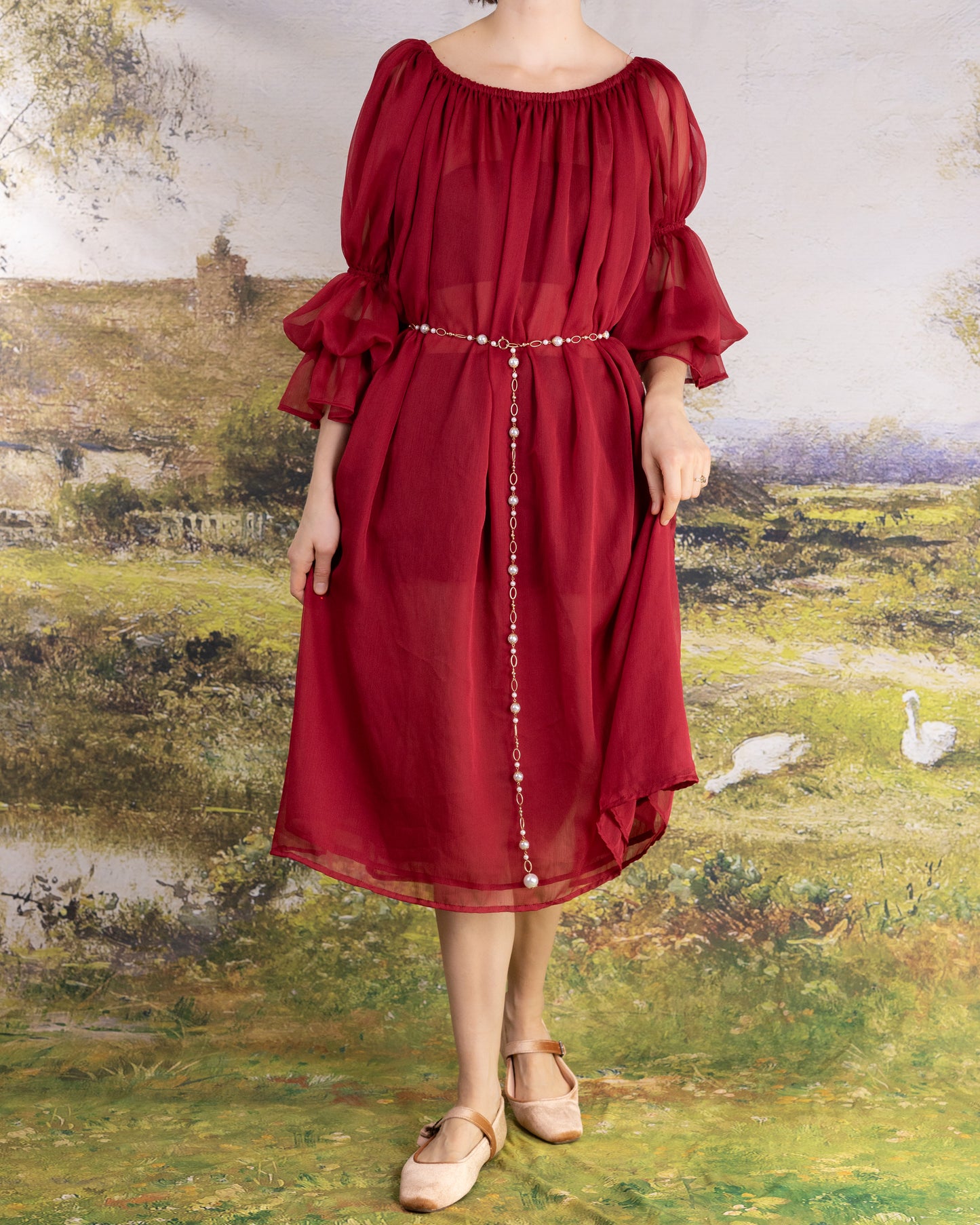 Woman wearing a flowing Red Chiffon Chemise with puffed sleeves and a delicate pearl belt standing in front of a countryside backdrop