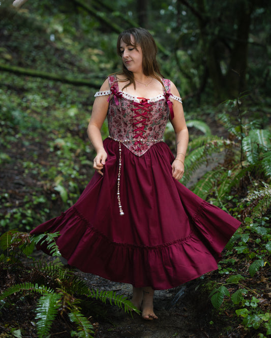 Woman wearing a Raspberry Rose Jacquard Corset twirling in a forest with a flowing burgundy skirt and delicate straps