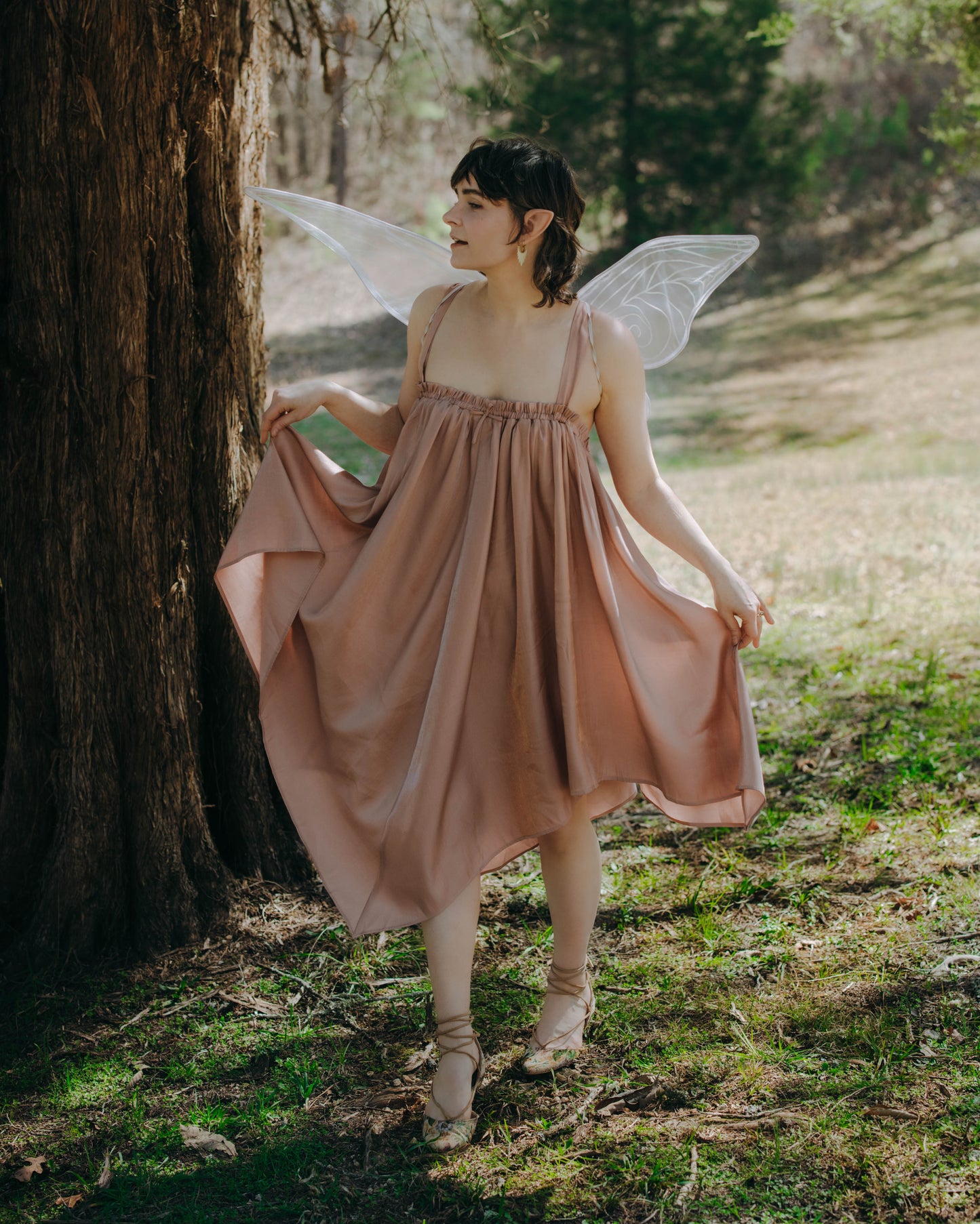 Woman wearing a light pink flowing Flutter Fairy Dress with adjustable straps standing by a tree in a forest setting.