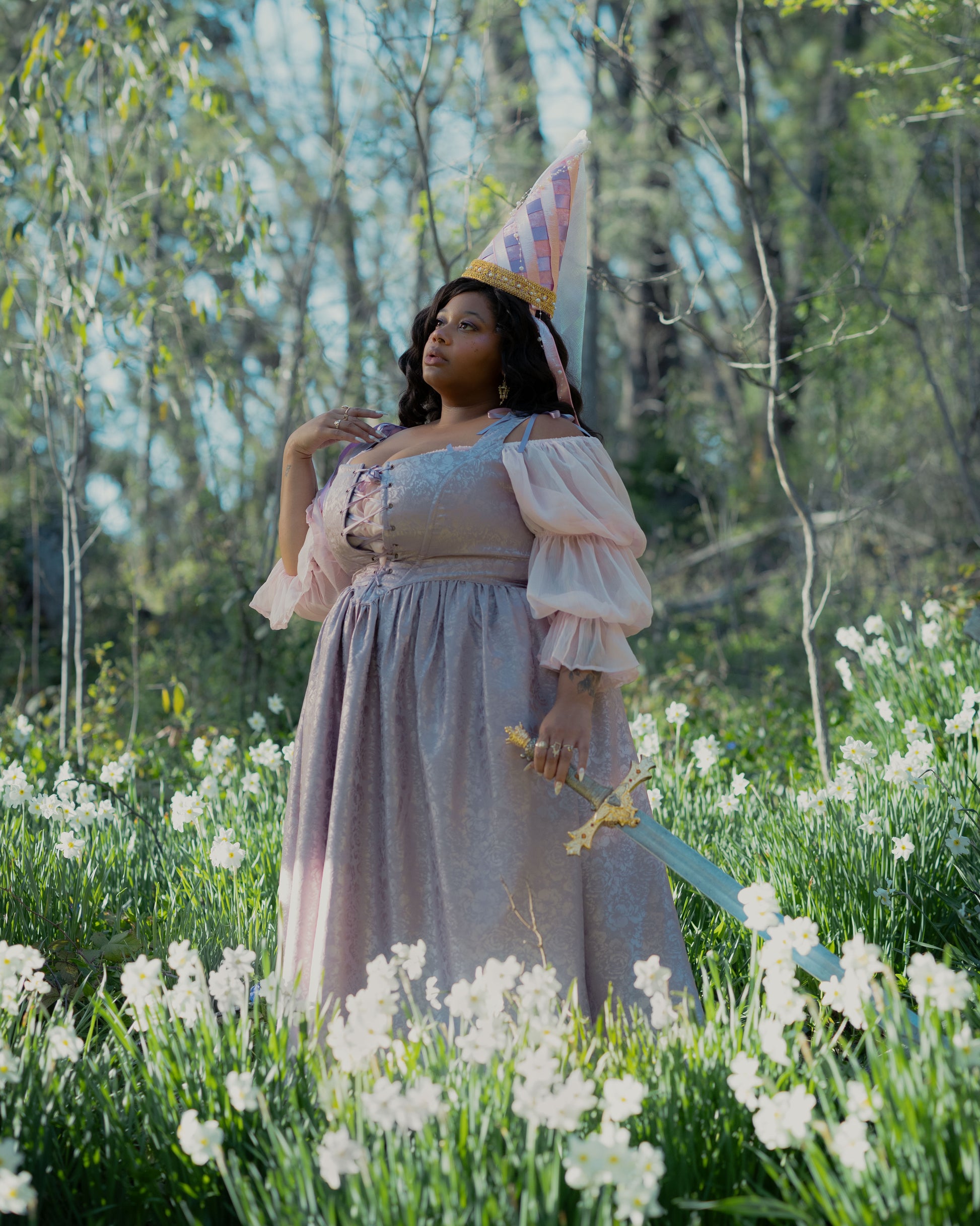 Woman in lavender satin corset dress with puff sleeves and cone hat holding a sword in a flower-filled forest.