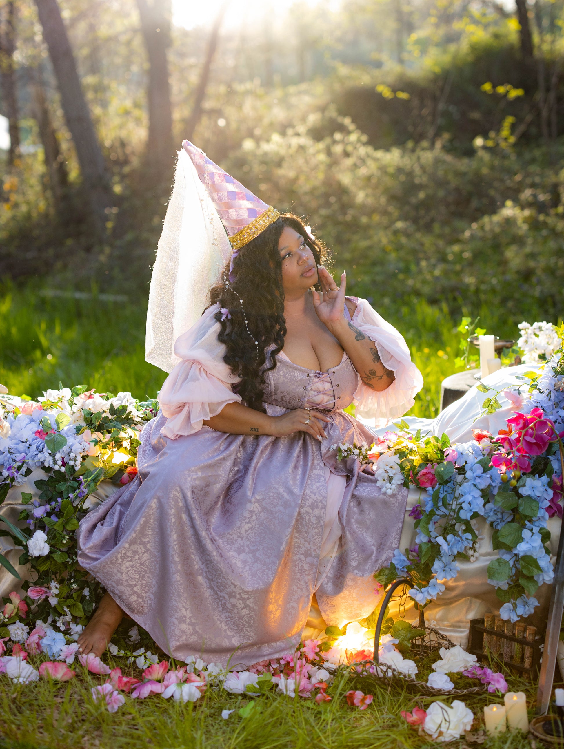 Woman in lavender floral satin corset dress with puff sleeves and cone hat sitting among colorful flowers outdoors