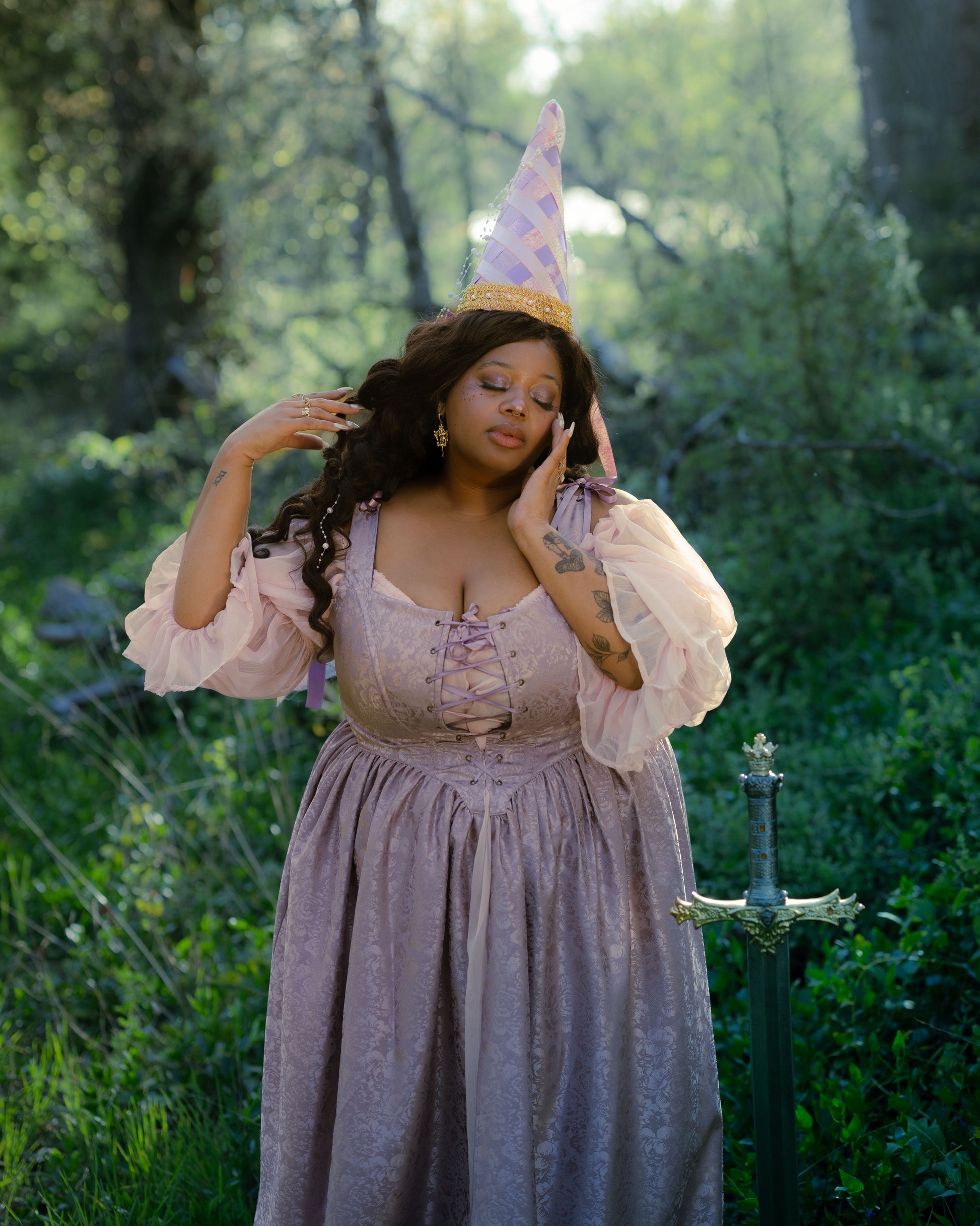 Woman in lavender satin corset dress with puff sleeves and pointed hat stands in enchanted forest near sword.