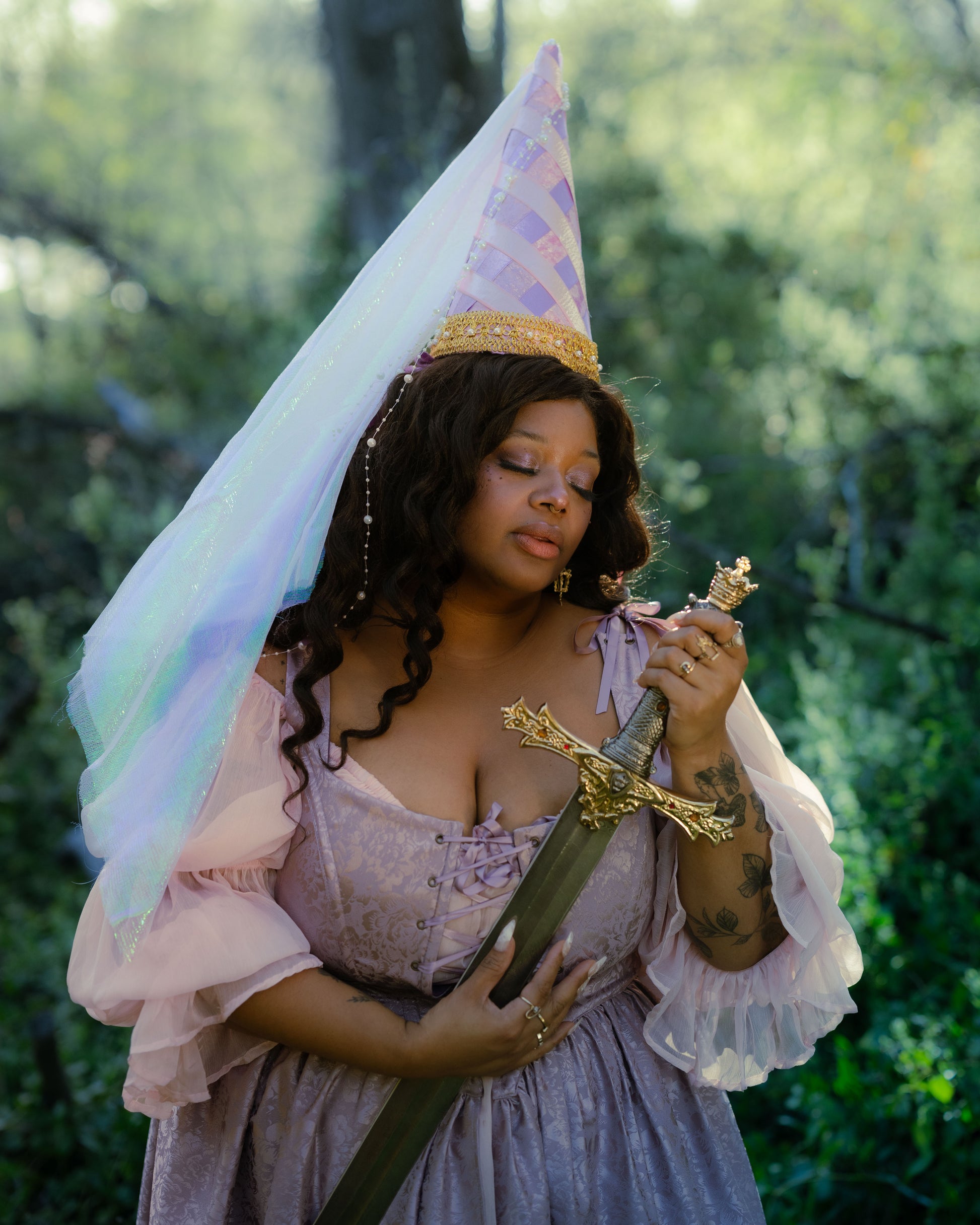 Woman wearing a lavender satin floral corset dress holding an ornate sword in a forest setting with a fairy tale hat.
