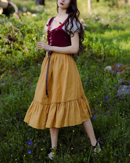 Woman wearing dark red velvet corset top with mustard skirt standing in a green field holding flowers