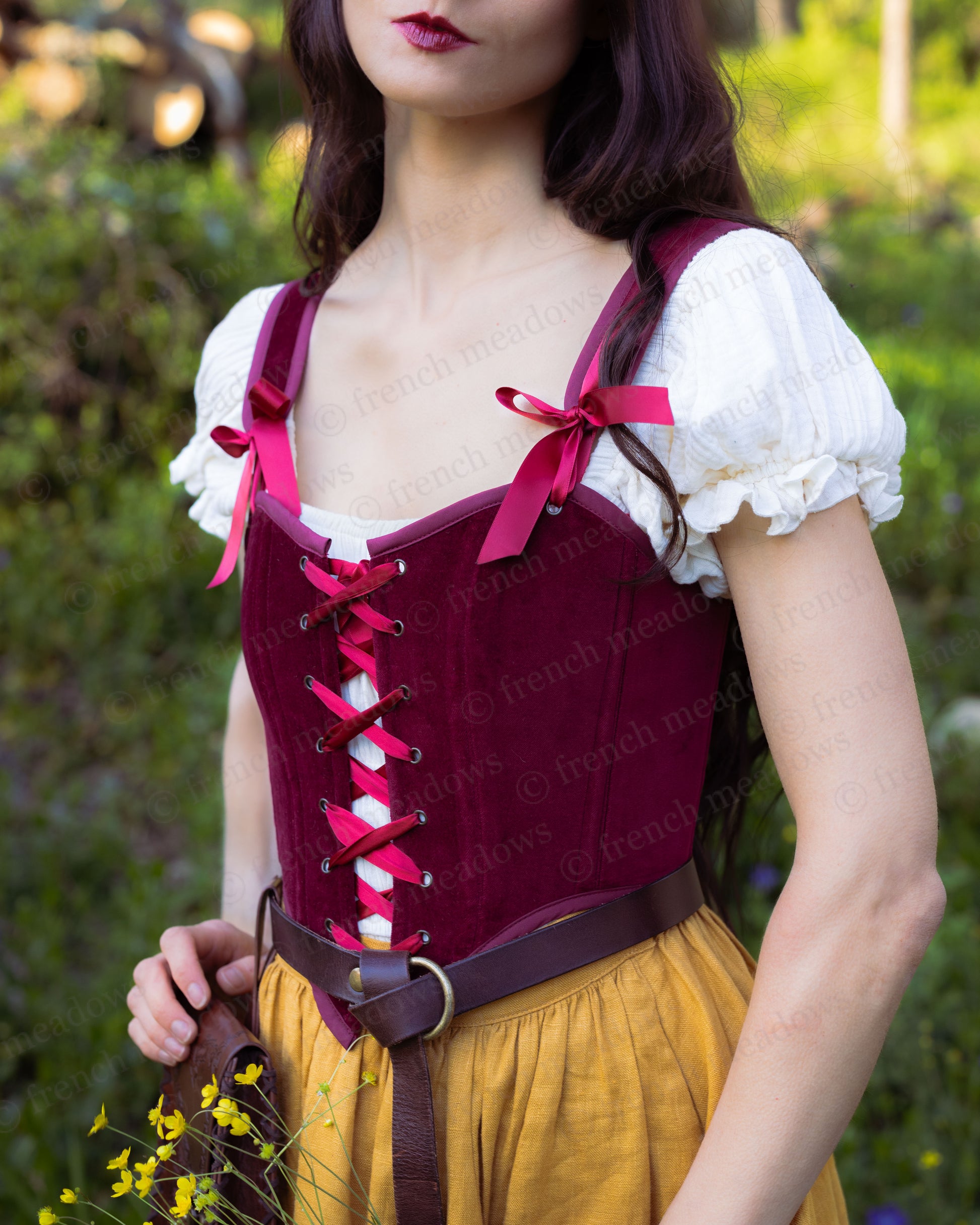 Woman wearing a dark red velvet corset top with lace-up front and ribbons over a white blouse outdoors.