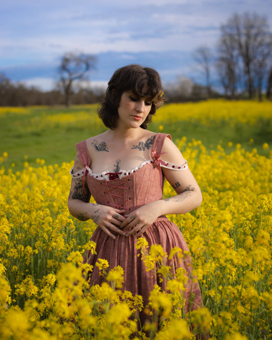 Young woman with tattoos wearing Strawberry Floral Regency Stays dress standing in yellow flower field under blue sky
