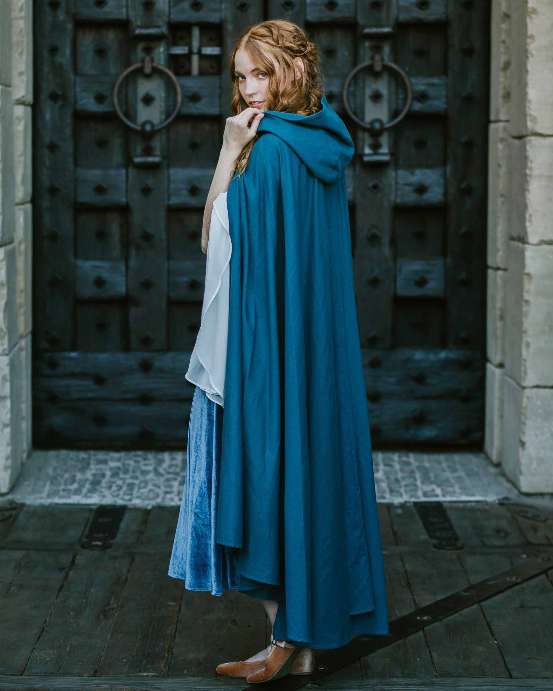 Woman wearing a blue linen renaissance cloak with hood standing in front of a large wooden door