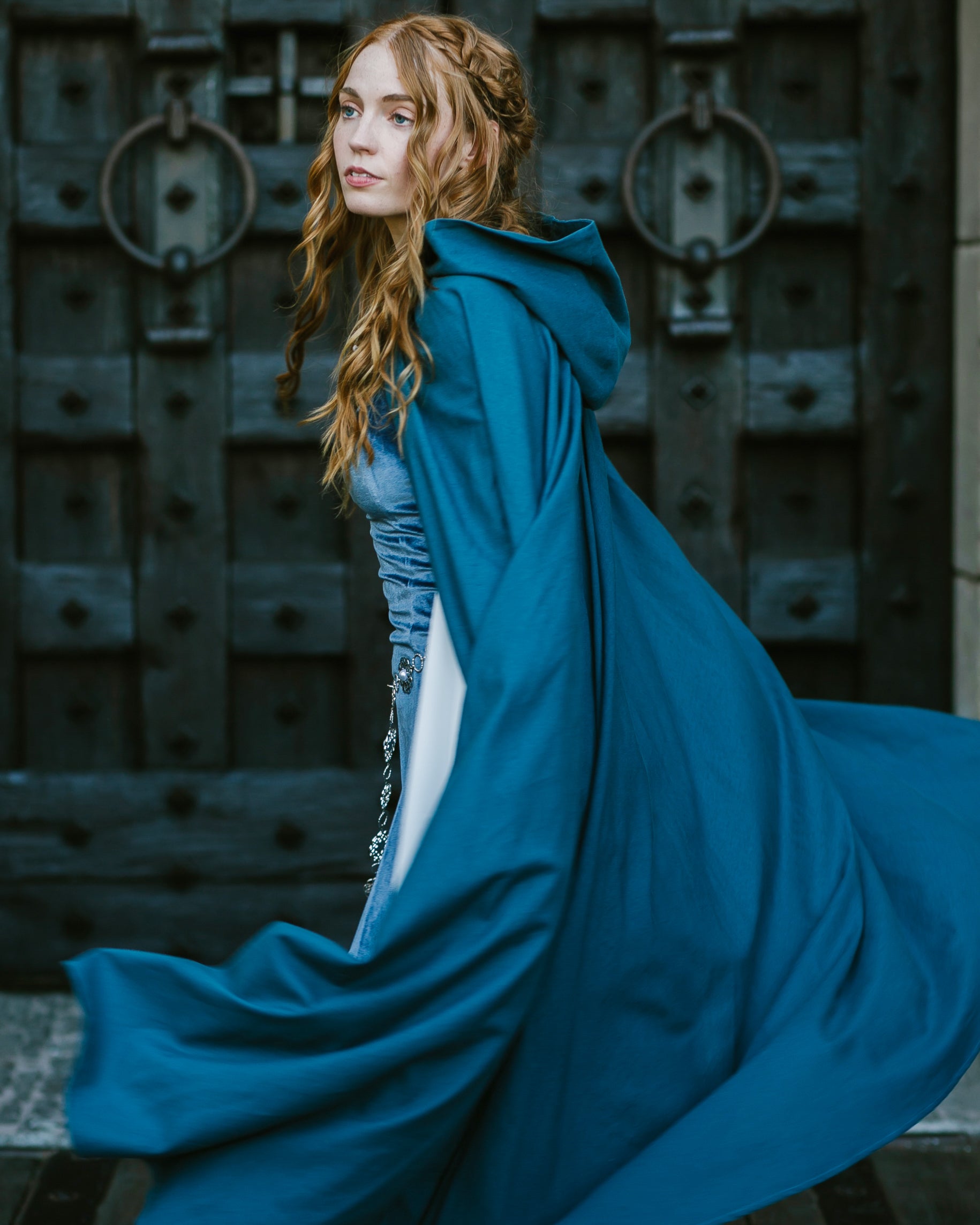 Woman wearing a flowing blue linen renaissance cloak with hood standing in front of large wooden doors