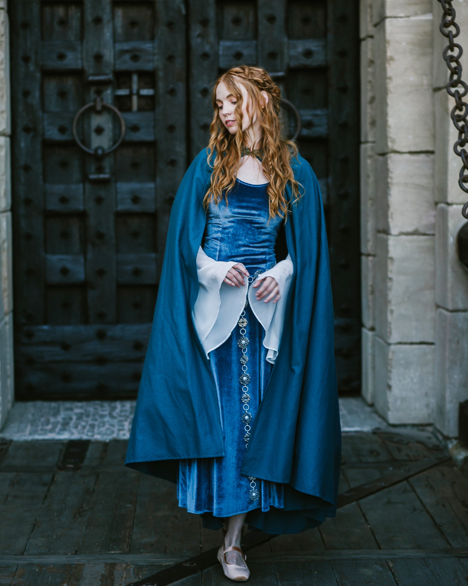 Woman wearing a blue Linen Renaissance Cloak over a velvet medieval dress standing in front of a large wooden door