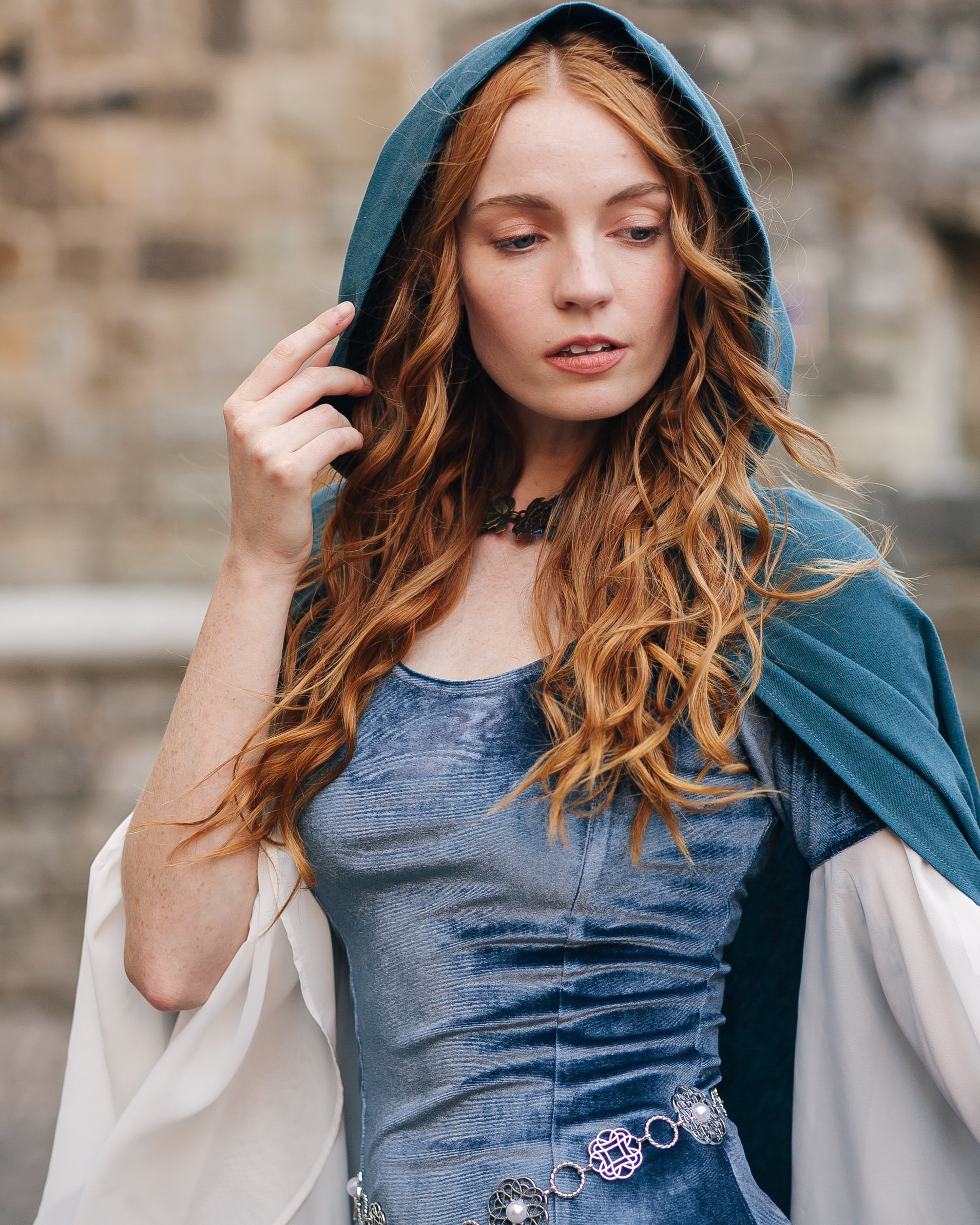 Woman with long red hair wearing a blue linen renaissance cloak with hood over a velvet dress by a stone wall