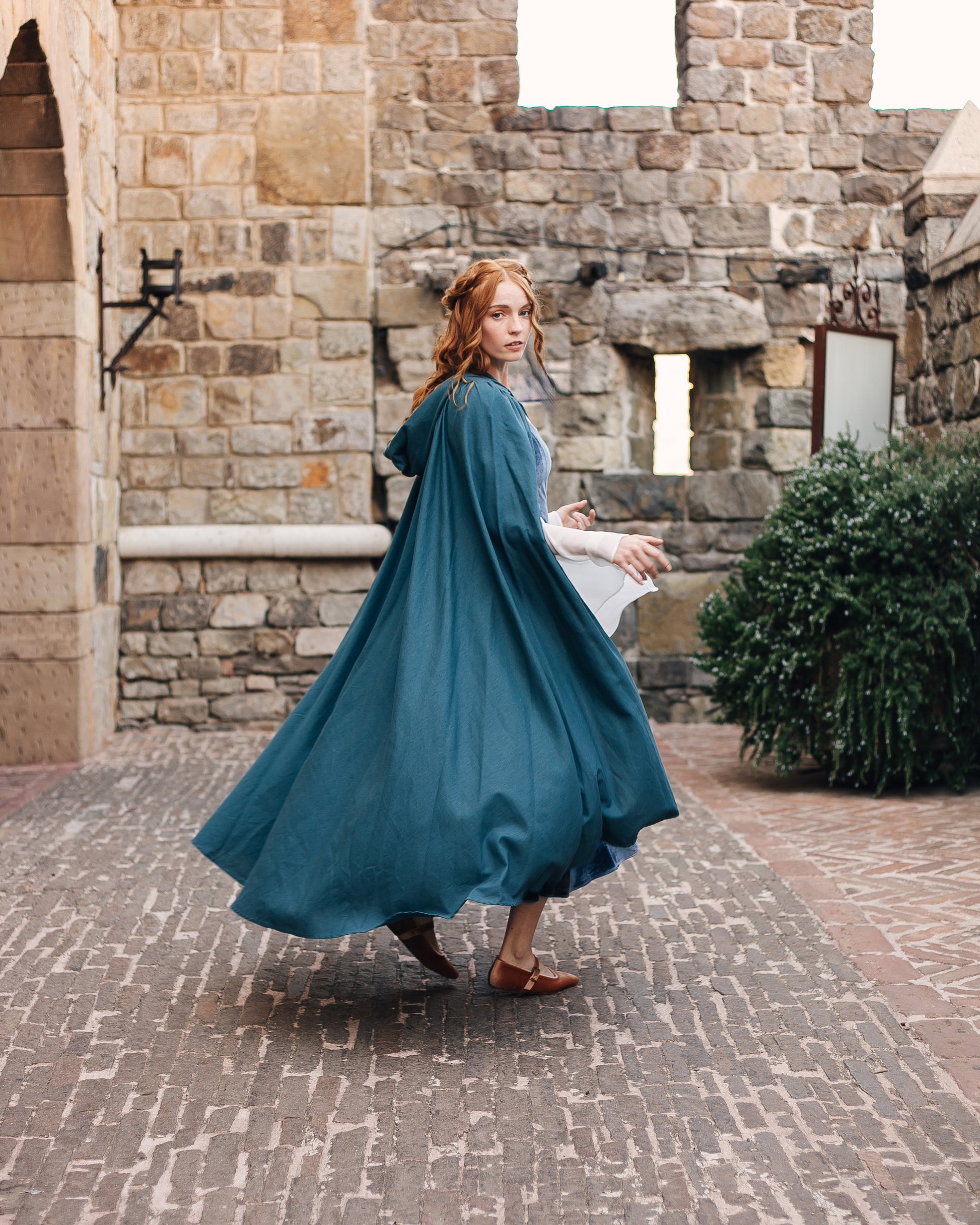 Woman wearing a flowing blue Linen Renaissance Cloak walking in a stone courtyard with medieval architecture background.