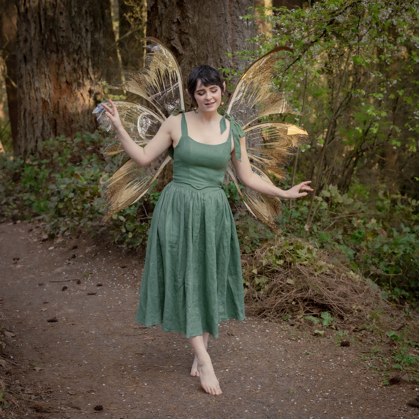 Woman wearing an ivy green linen midi skirt with fairy wings in a forest setting, barefoot and smiling.