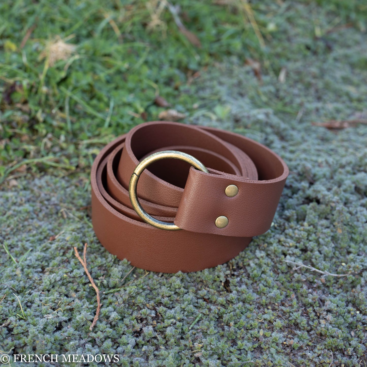 Brown vegan leather renaissance belt with brass ring buckle and rivets displayed on mossy ground